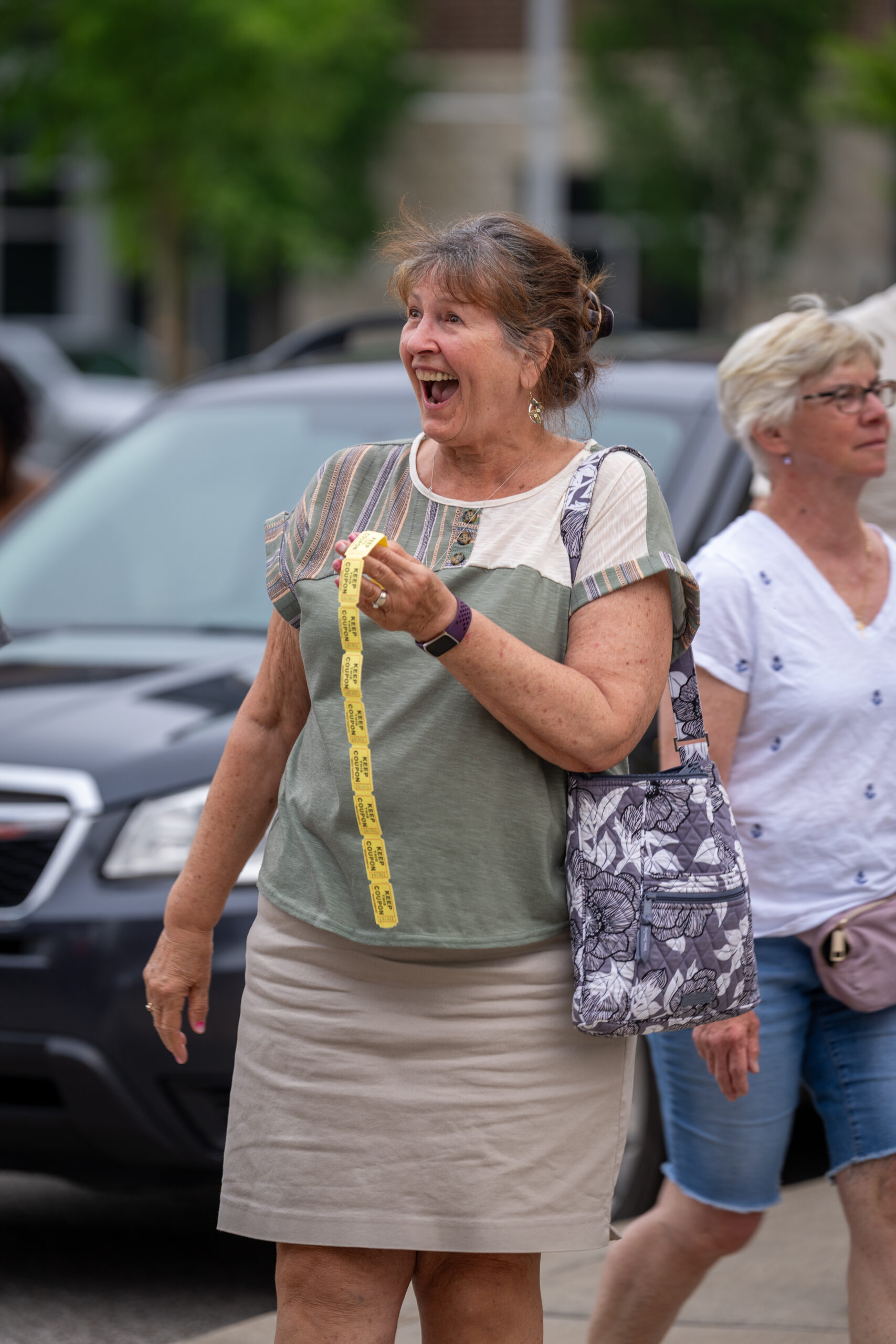 A smiling woman stands outdoors at Sunset Music Fest holding a strip of yellow tickets, wearing a green and beige top and tan skirt with a patterned bag. Another woman in glasses walks behind her. Cars and trees are in the background.