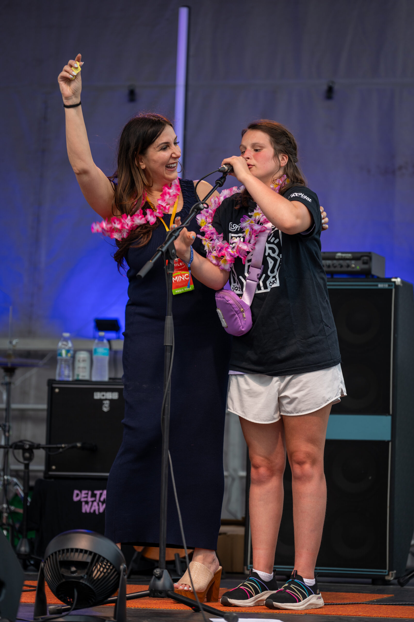 Two women stand on stage at Sunset Music Fest, both wearing pink leis. One in a dark dress raises her hand, smiling and holding a microphone, while the other, in a black T-shirt and white shorts, speaks into the mic.
