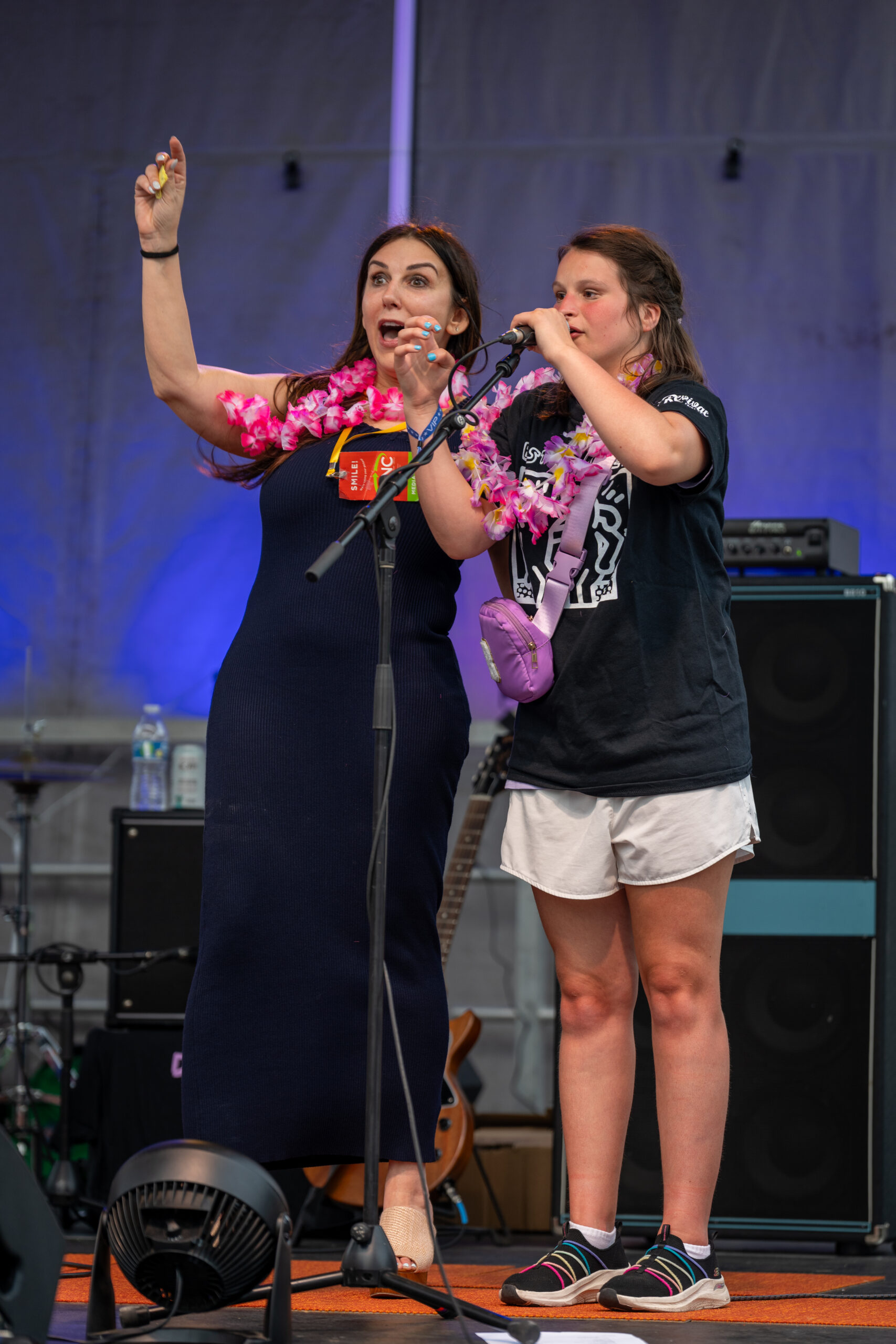 Two women stand on stage at Canonsburg Live Music; one in a navy dress gestures as she speaks, while the other, in a black T-shirt and white shorts, holds a microphone. Both wear colorful leis, with music equipment visible in the background.