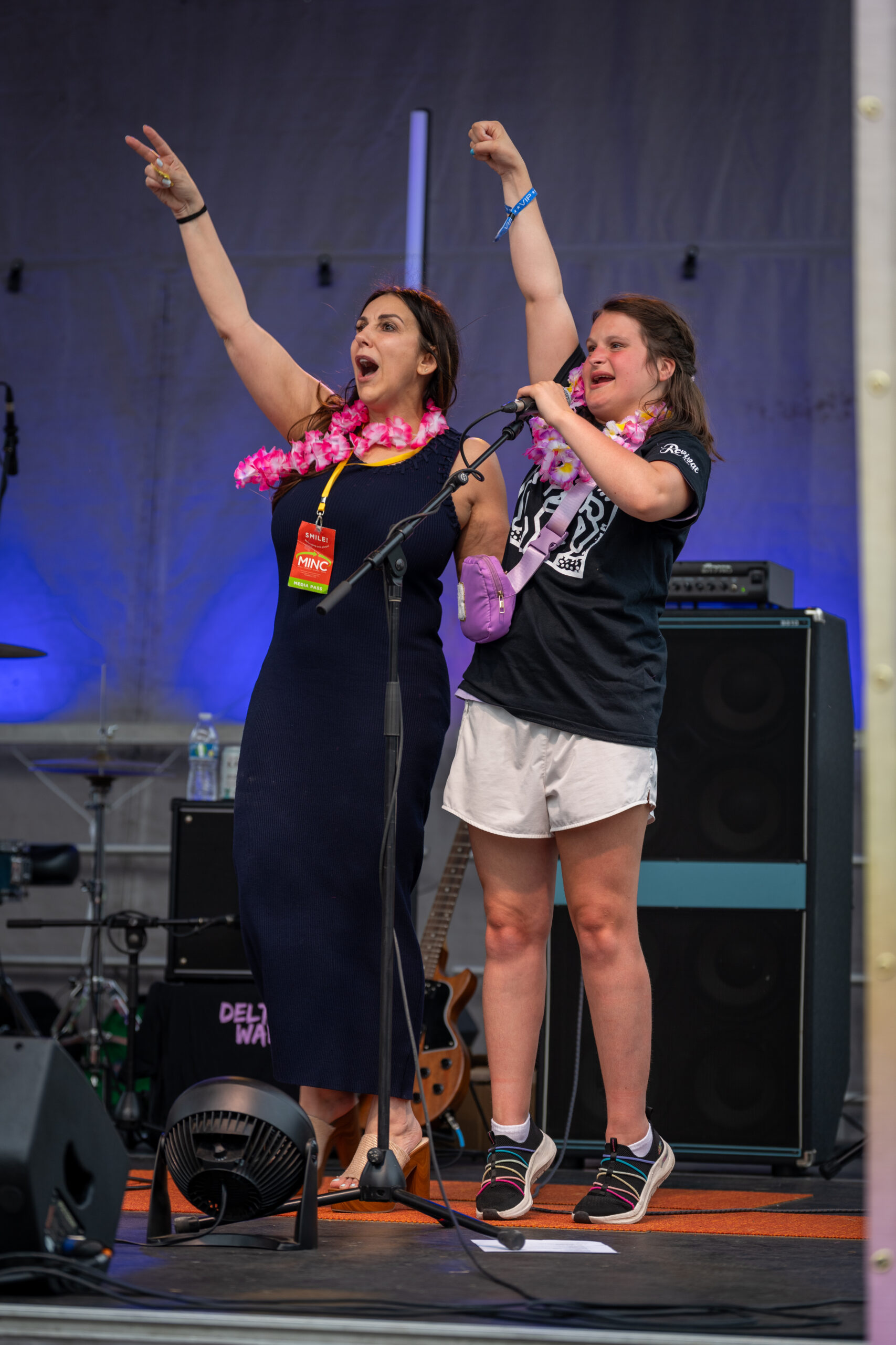 Two women onstage at Sunset Music Fest, both wearing flower leis, smile and raise their arms enthusiastically. One holds a microphone, with musical equipment, a guitar, and speakers visible behind them at this lively Canonsburg live music event.