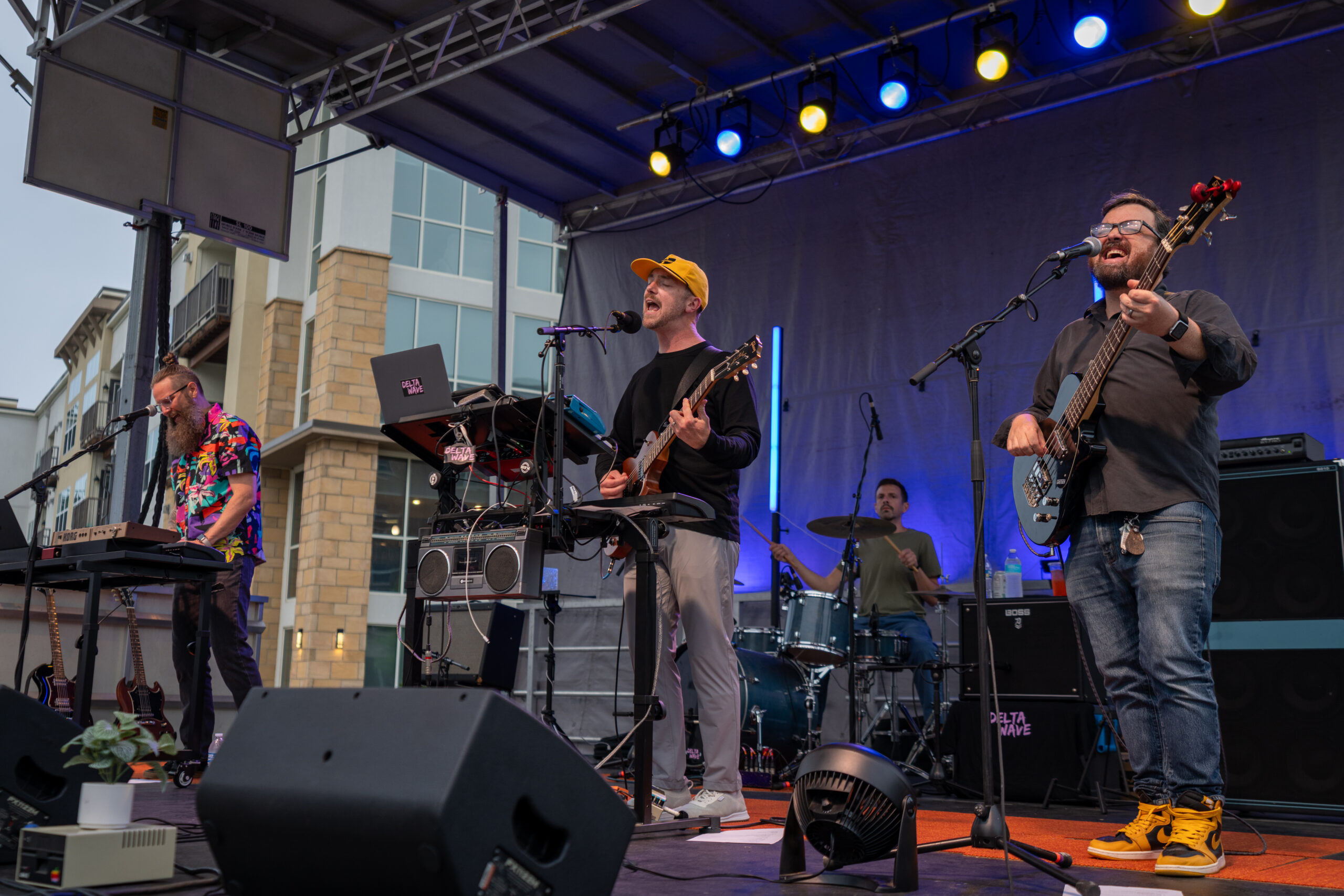A four-member band performs energetically on an outdoor stage at Sunset Music Fest; one member plays keyboard, another guitar and sings, a third plays drums, and the fourth plays bass guitar, all under colorful stage lights.