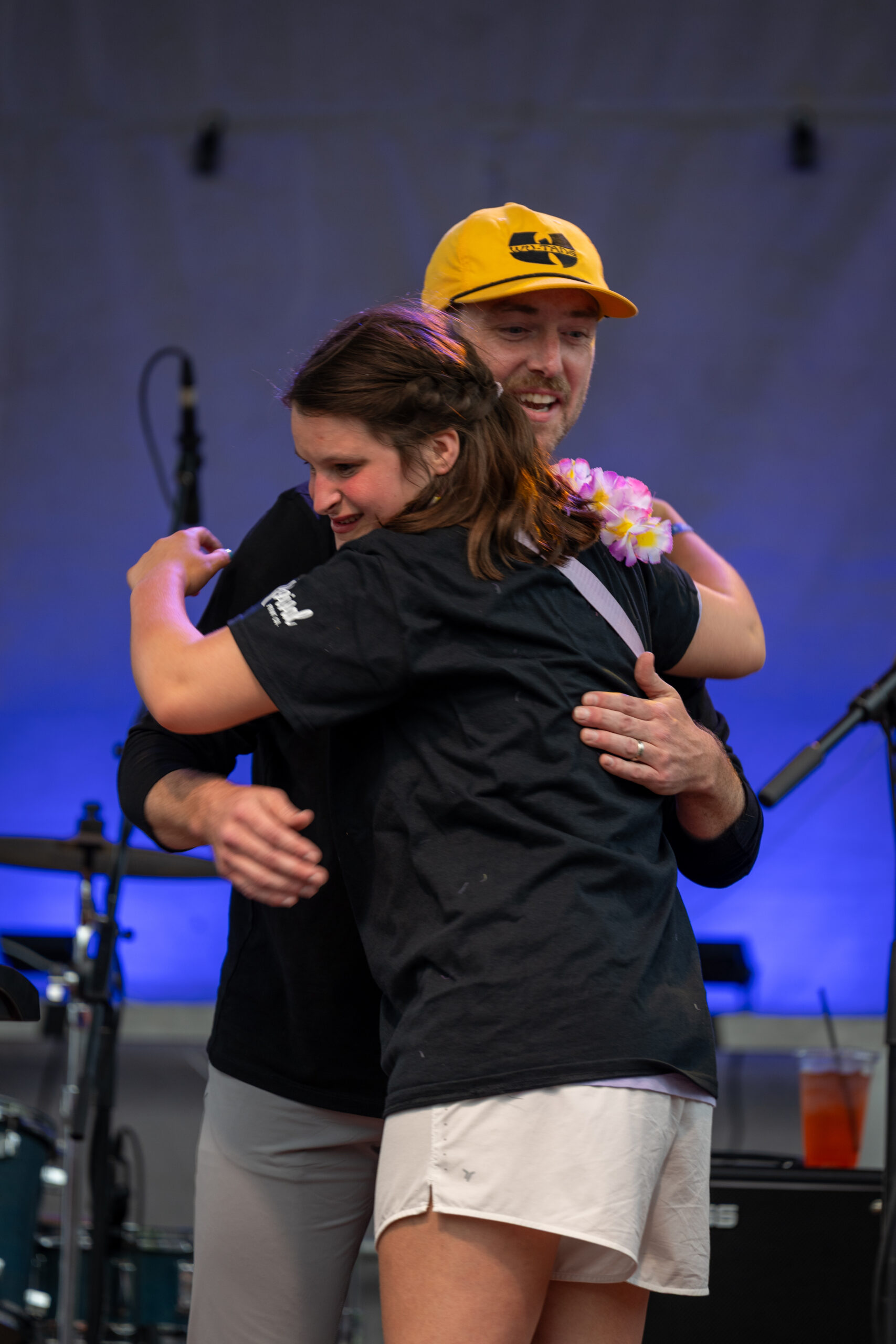 A woman and a man warmly hug on stage at the Sunset Music Fest. The man wears a yellow hat and lei, while the woman sports a black shirt and white shorts. Musical equipment and a drink are visible in the background.