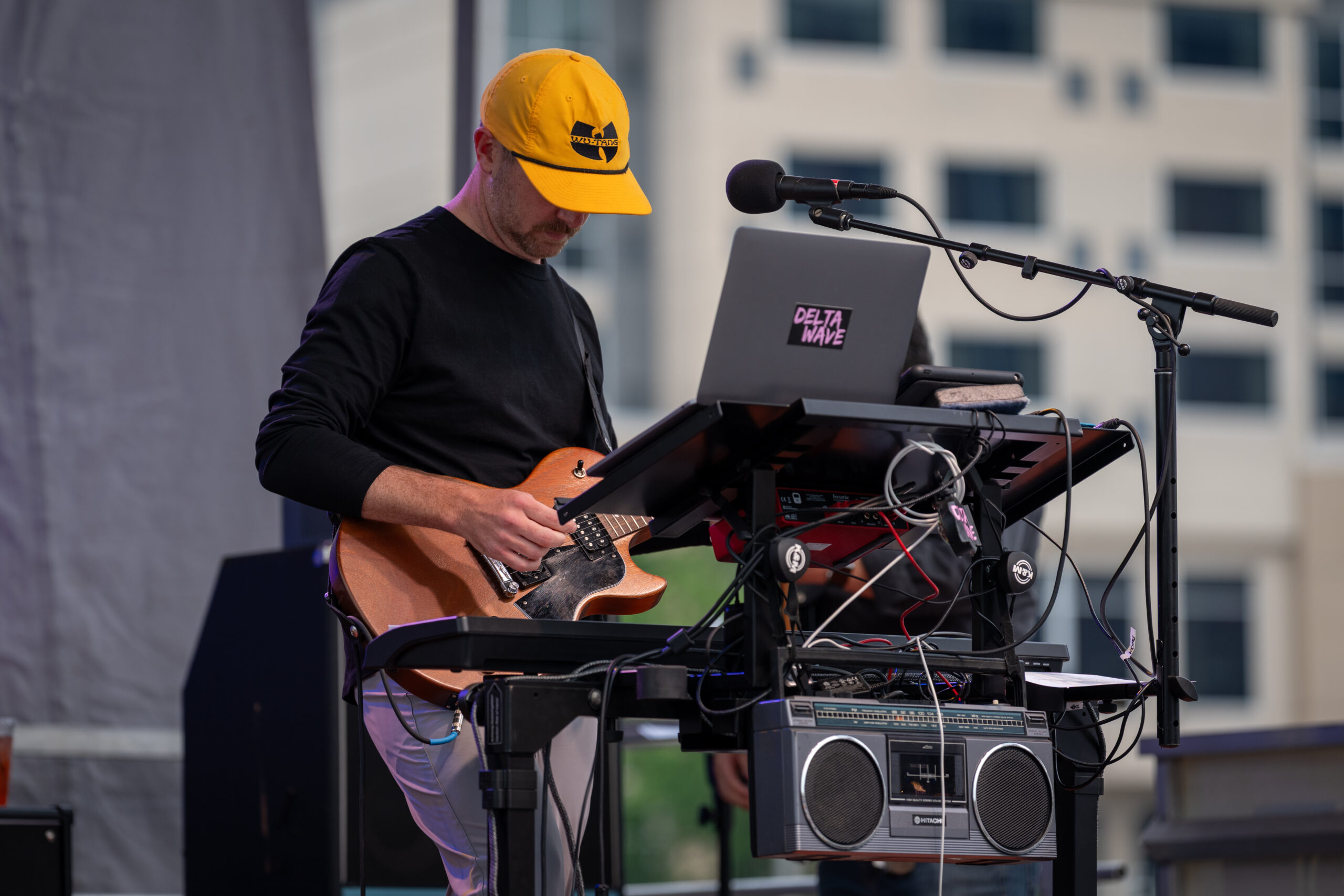 A musician in a yellow cap plays an electric guitar at a keyboard and laptop setup on an outdoor stage during Sunset Music Fest, with audio equipment and city buildings in the background.