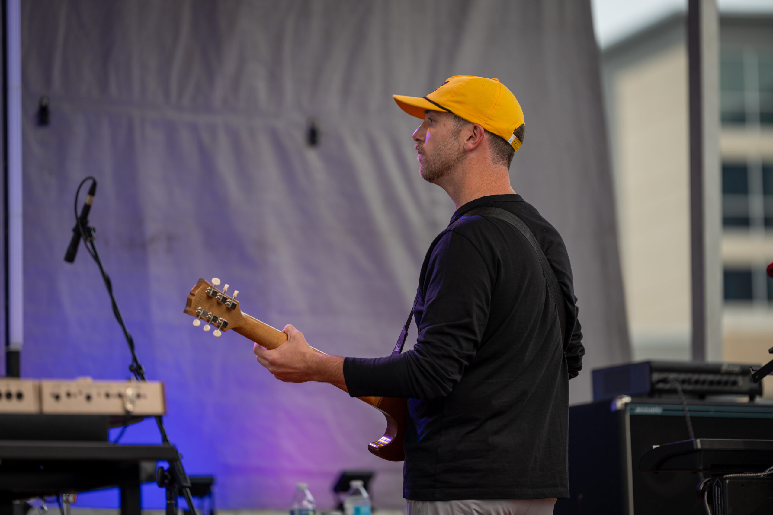 A man wearing a yellow cap and black long-sleeve shirt plays an electric guitar on an outdoor stage at Sunset Music Fest, with blurred musical equipment and a building in the background.
