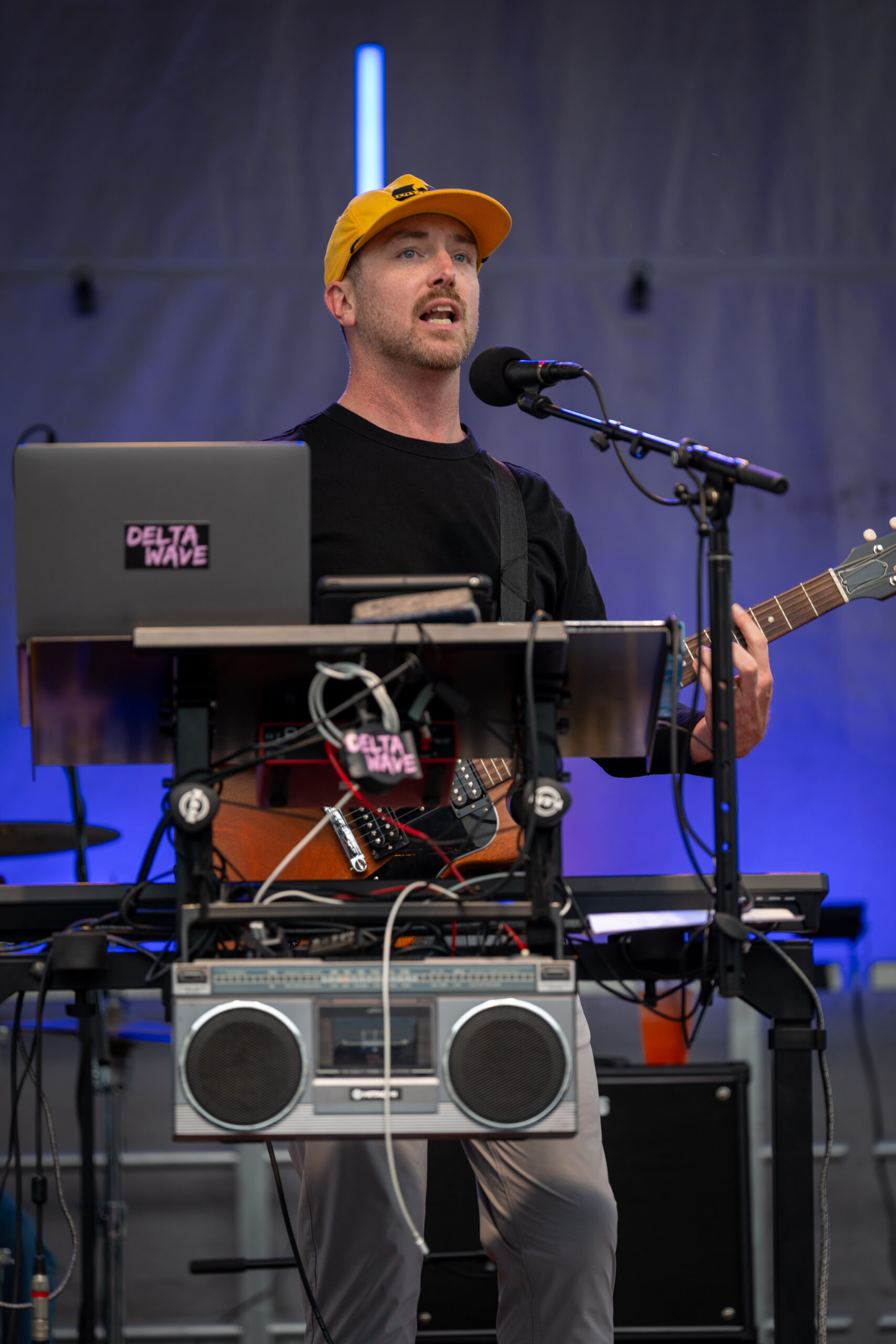 A musician wearing a yellow cap sings into a microphone while playing an electric guitar. He stands behind a keyboard setup with wires, a laptop, and a boombox, performing on an outdoor stage at the Sunset Music Fest.