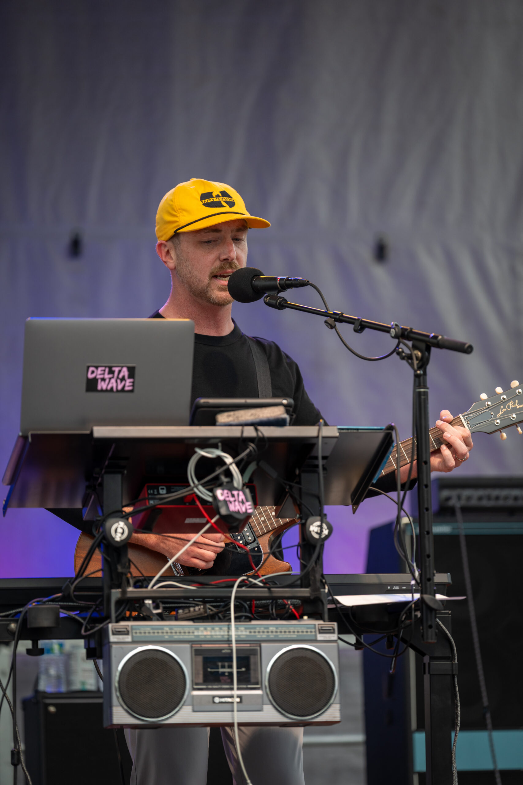 A musician wearing a yellow hat sings into a microphone while playing an electric guitar. He stands behind a stand with a laptop, audio equipment, and cables on an outdoor stage at Canonsburg Live Music.