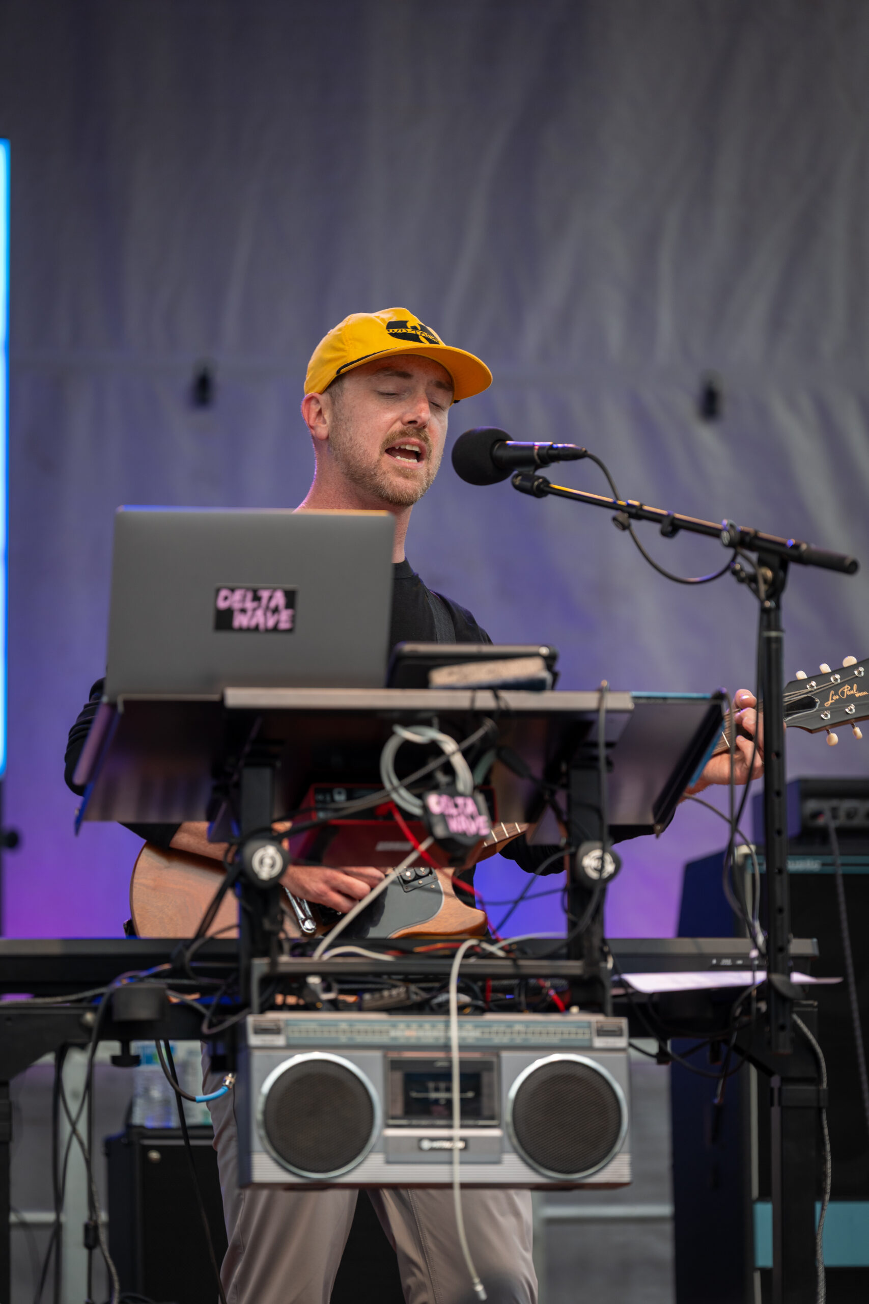 A musician wearing a yellow cap sings into a microphone while playing guitar, surrounded by electronic equipment and a keyboard on stage at Canonsburg Live Music. A boombox sits on a stand in front of him.