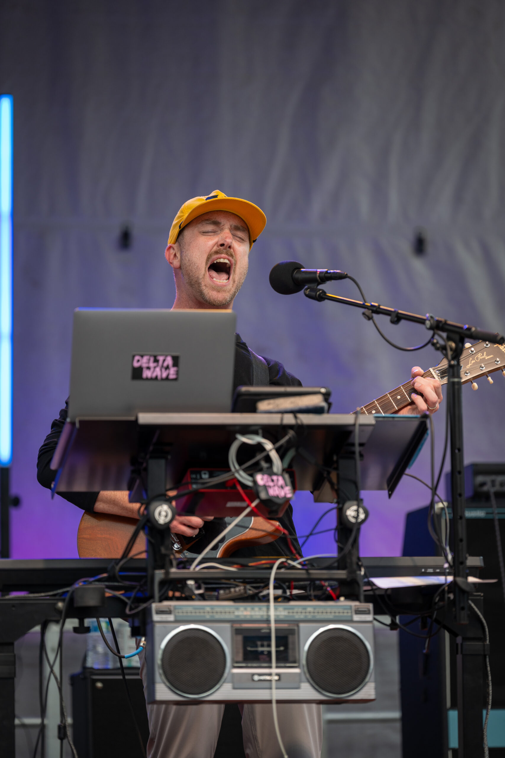 A musician wearing a yellow cap passionately sings and plays guitar on stage behind a laptop labeled “DELTA WAVE” at Sunset Music Fest, with a microphone, music equipment, and a visible boombox in front of him.