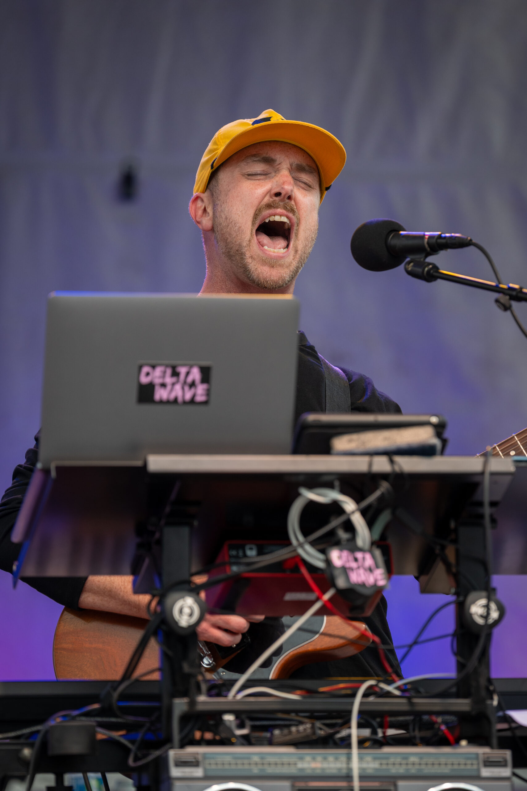 A musician wearing a yellow cap passionately sings into a microphone while playing guitar, standing behind electronic audio equipment labeled DELTA WAVE during a live performance at Sunset Music Fest.