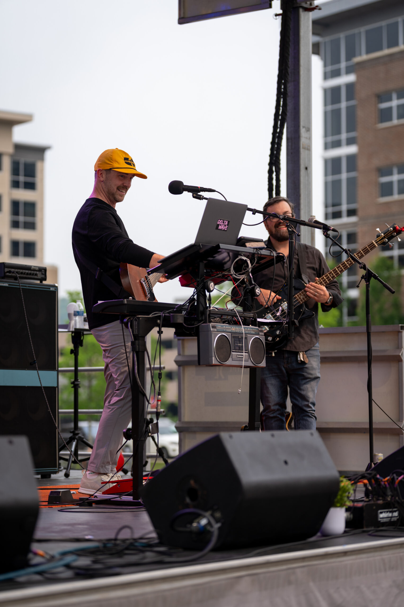 Two musicians perform on an outdoor stage at Sunset Music Fest; one wears a yellow cap and plays keyboard and guitar, while the other plays guitar and sings into a microphone. Buildings are visible in the background.