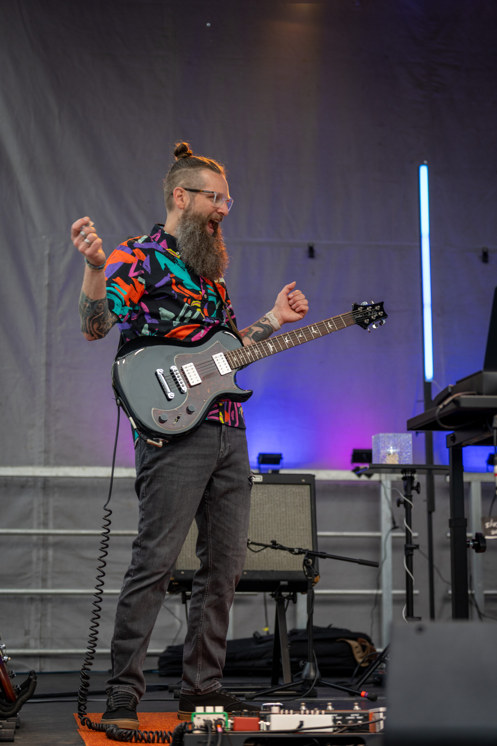 A bearded man with glasses, wearing a colorful shirt, plays an electric guitar on stage at the Sunset Music Fest. He is smiling and standing near music equipment, with purple and blue lights in the background.