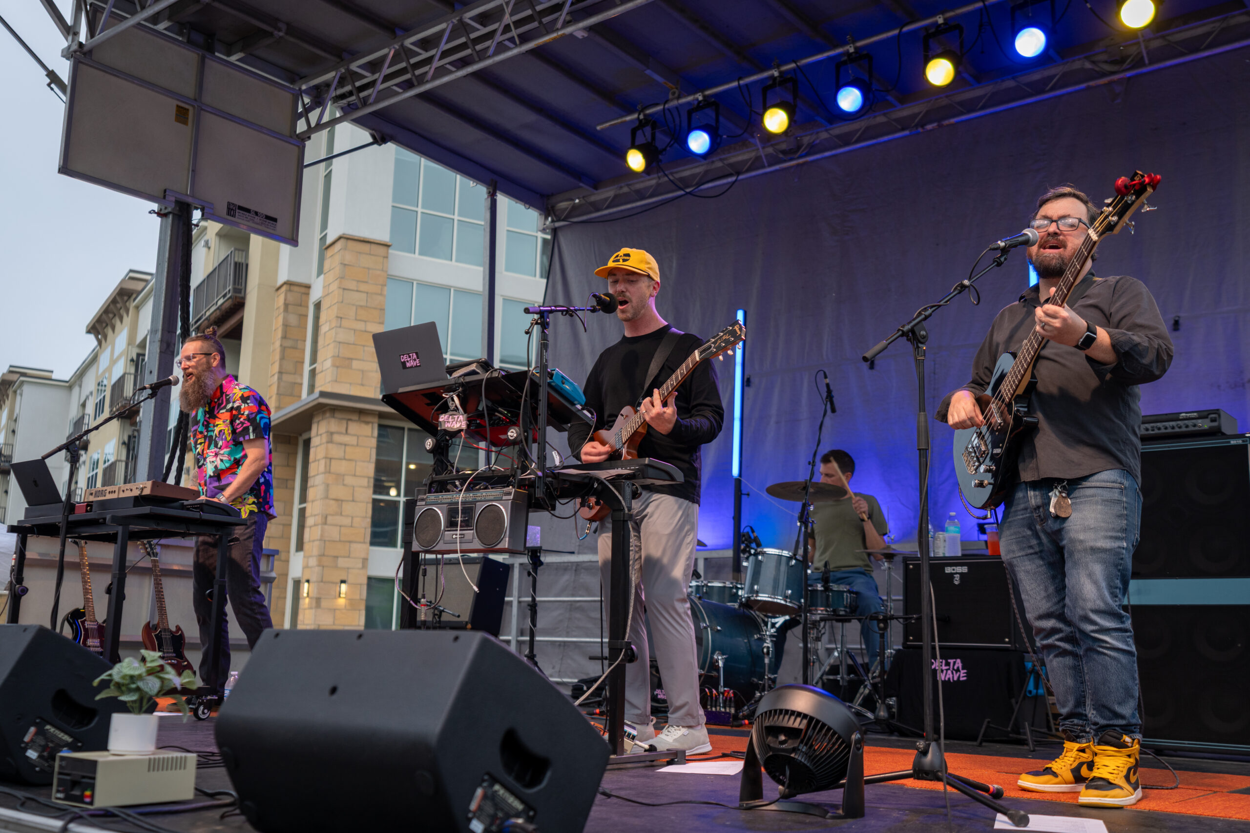 A four-member band performs on an outdoor stage at the Sunset Music Fest, playing keyboard, guitars, and drums under colorful lights. Modern buildings are visible in the background.