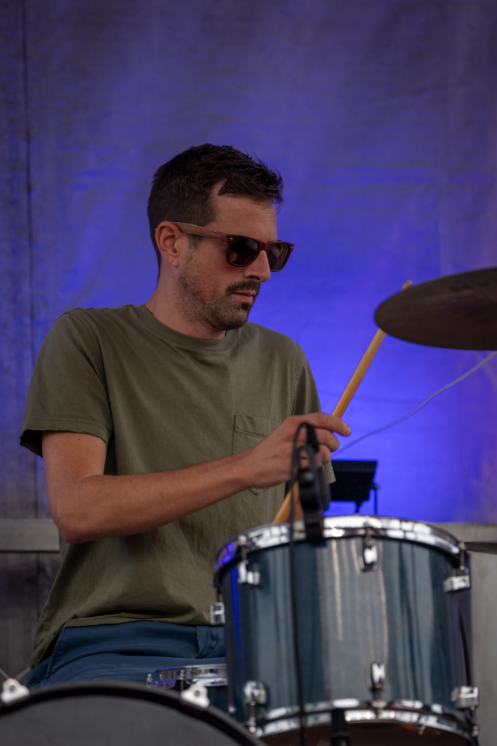 A man wearing sunglasses and a green t-shirt plays a drum set on stage at the Sunset Music Fest, with a blue-lit background behind him.