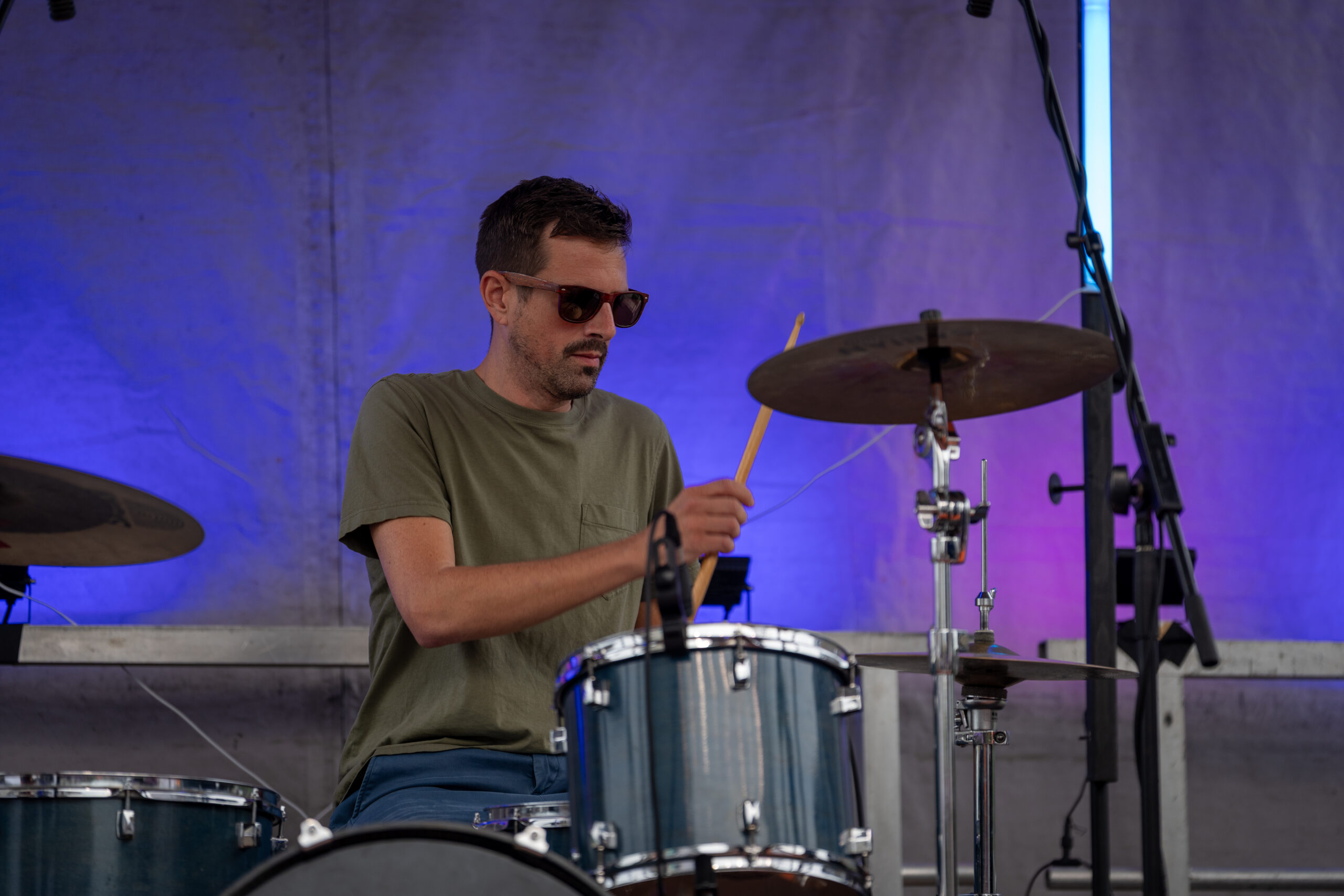 A man wearing sunglasses and a green t-shirt plays a drum set on an outdoor stage at Canonsburg Live Music, with purple and blue lighting in the background.