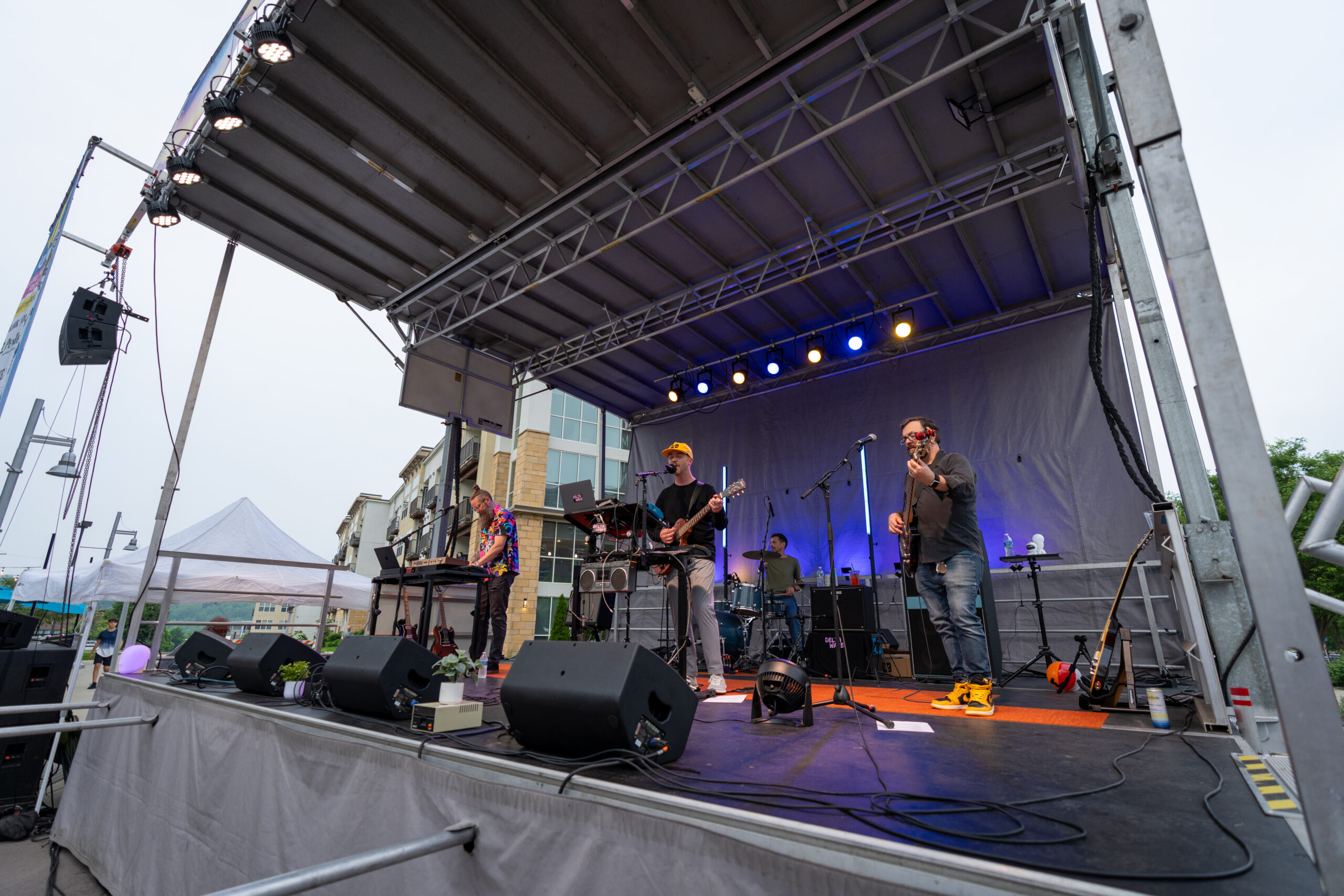 A band performs on an outdoor stage under a canopy of lights at Sunset Music Fest, with musicians playing guitar, keyboard, bass, and drums. Speakers and equipment fill the area as buildings rise in the background.