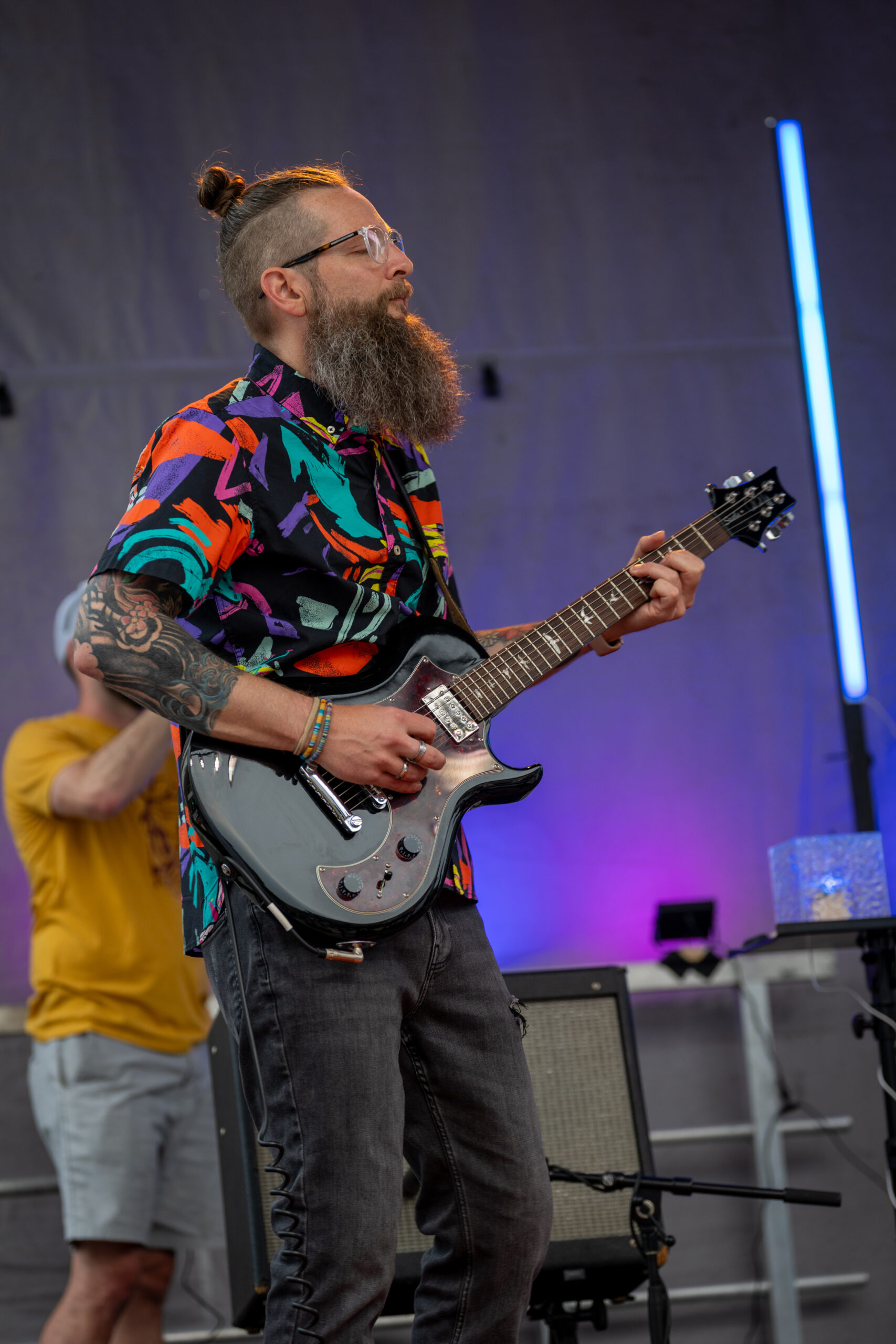 A bearded man with glasses, a colorful patterned shirt, and a man bun plays an electric guitar on stage at Sunset Music Fest. Purple and blue stage lights illuminate the background.