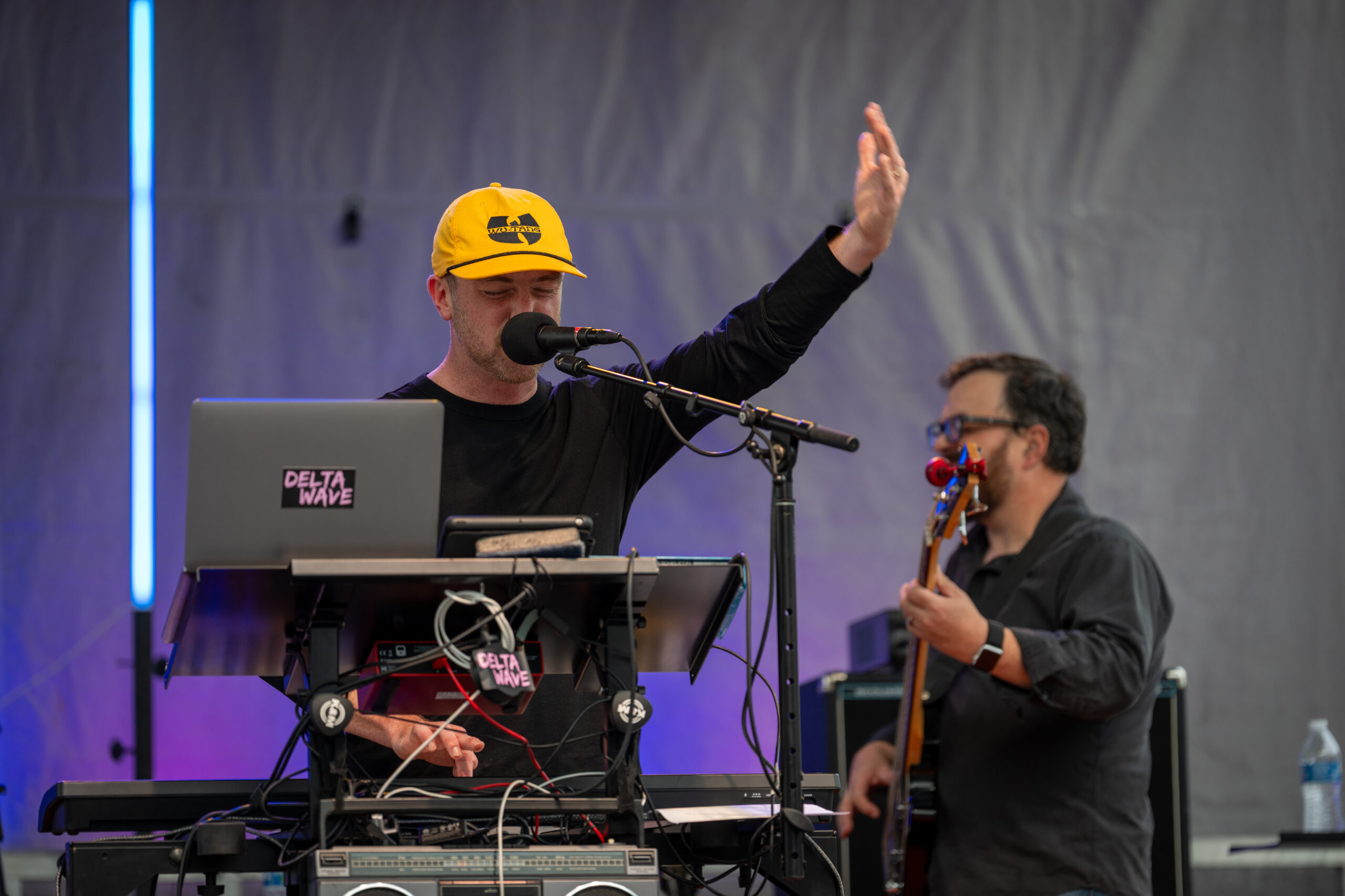 A musician wearing a yellow cap performs on stage with a microphone and electronic equipment at Sunset Music Fest, while another musician plays bass guitar in the background.