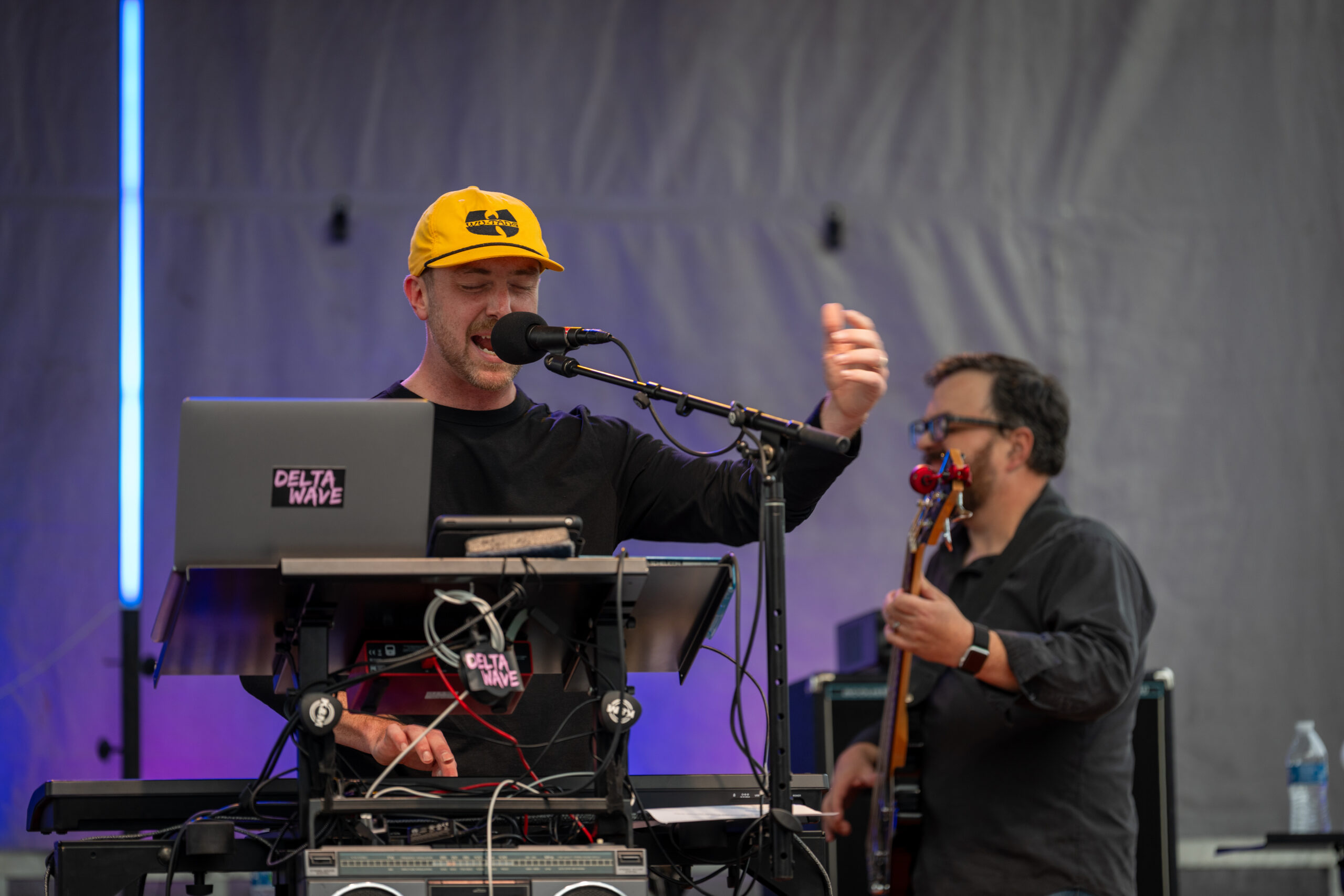 A musician in a yellow hat sings and plays keyboard on stage at Sunset Music Fest, while another musician in the background plays bass guitar. Both are surrounded by musical equipment with Delta Wave stickers.