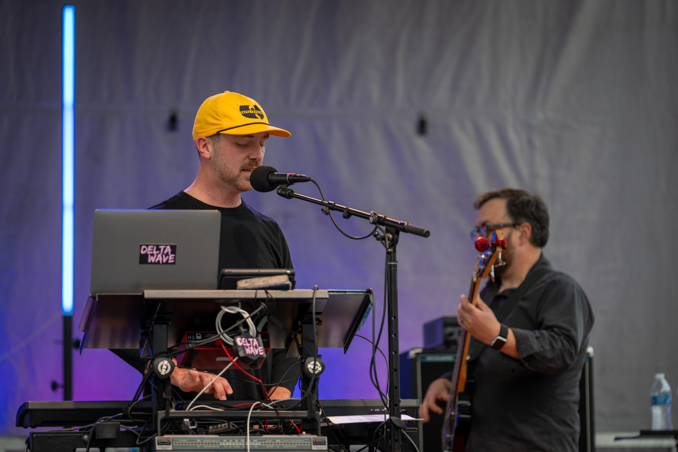 A musician wearing a yellow cap sings into a microphone while playing keyboards onstage at Sunset Music Fest. Another musician in the background plays bass guitar. A laptop with Delta Wave stickers is visible in the foreground.
