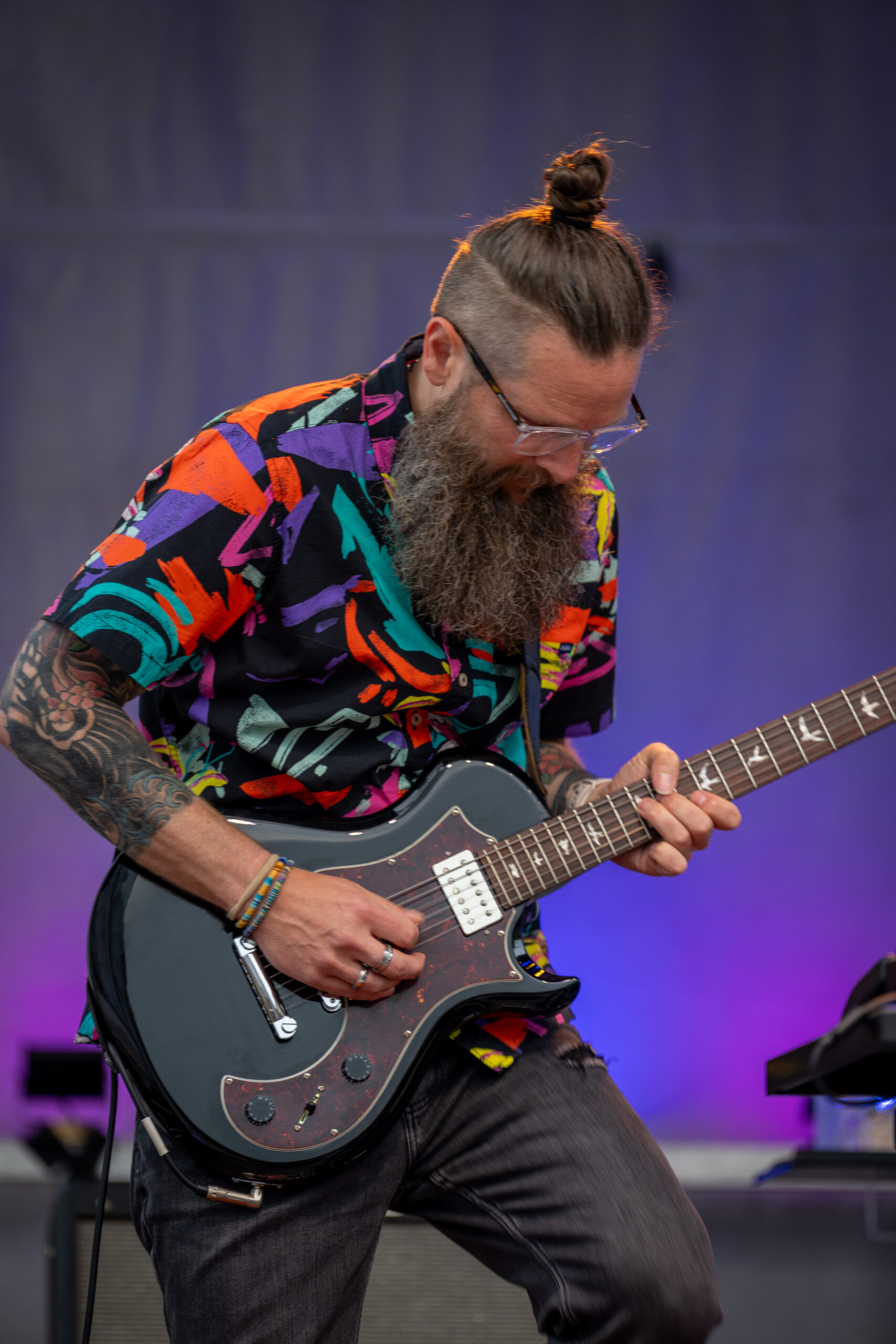 A bearded man with glasses, a bun, and tattoos plays electric guitar on stage at the Sunset Music Fest. He wears a colorful, patterned shirt and dark pants, with a purple-lit background behind him.