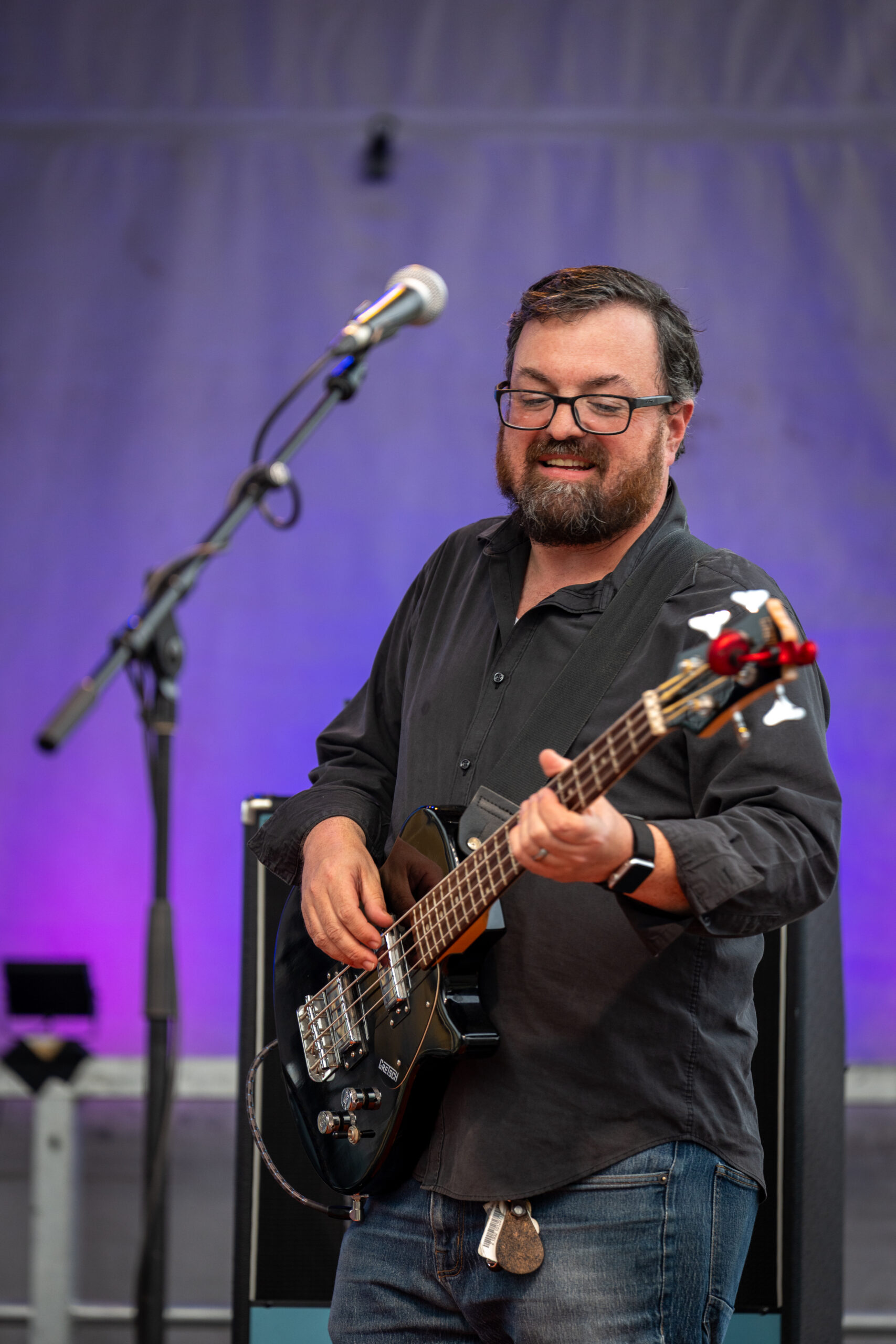A bearded man wearing glasses and a dark shirt plays an electric bass guitar on stage at Sunset Music Fest, smiling. A microphone stands in front of him as purple lights illuminate the background, capturing the joy of the music festival atmosphere.
