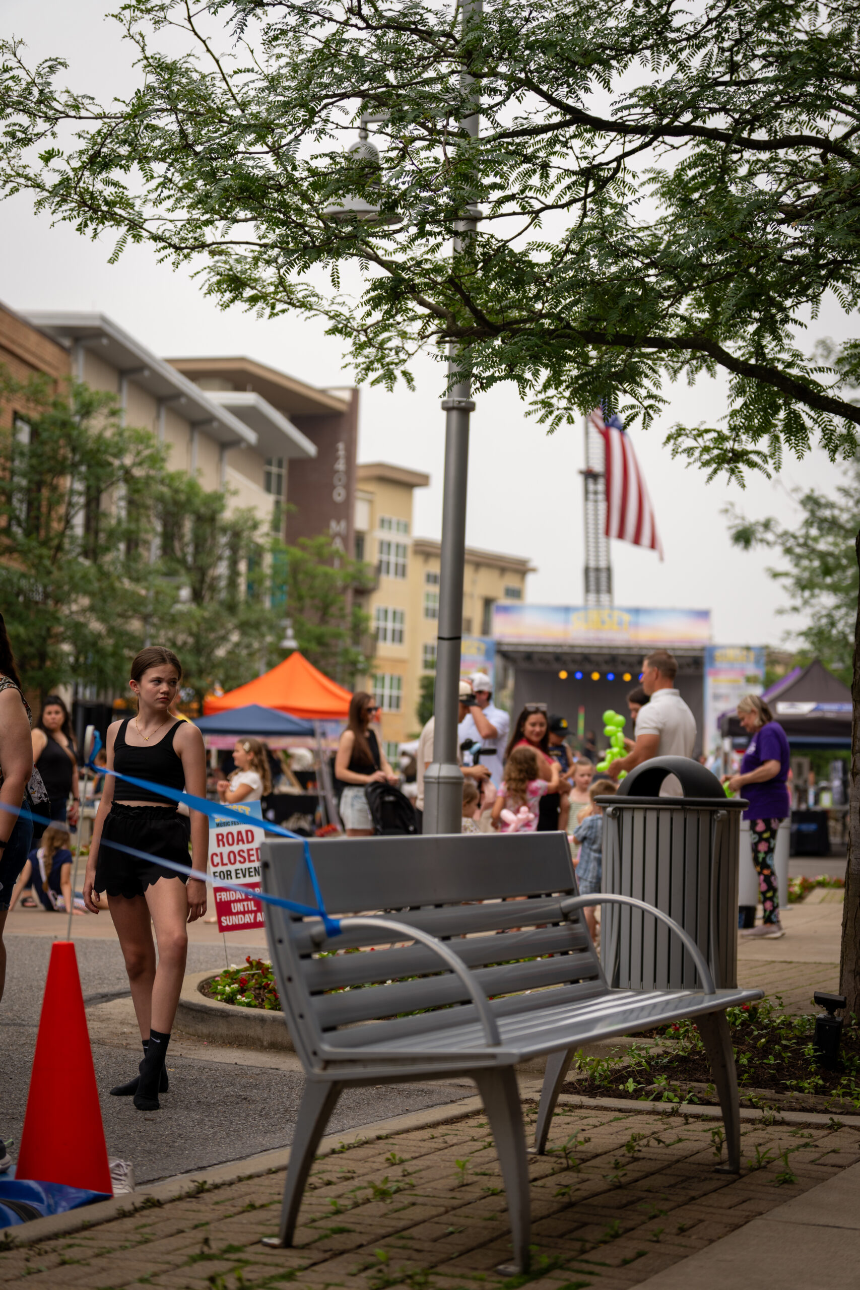 People gather at an outdoor street festival with tents, a stage, and an American flag in the background. A woman walks past a road closed sign near a bench and tree as Canonsburg Live Music brings energy to the scene.