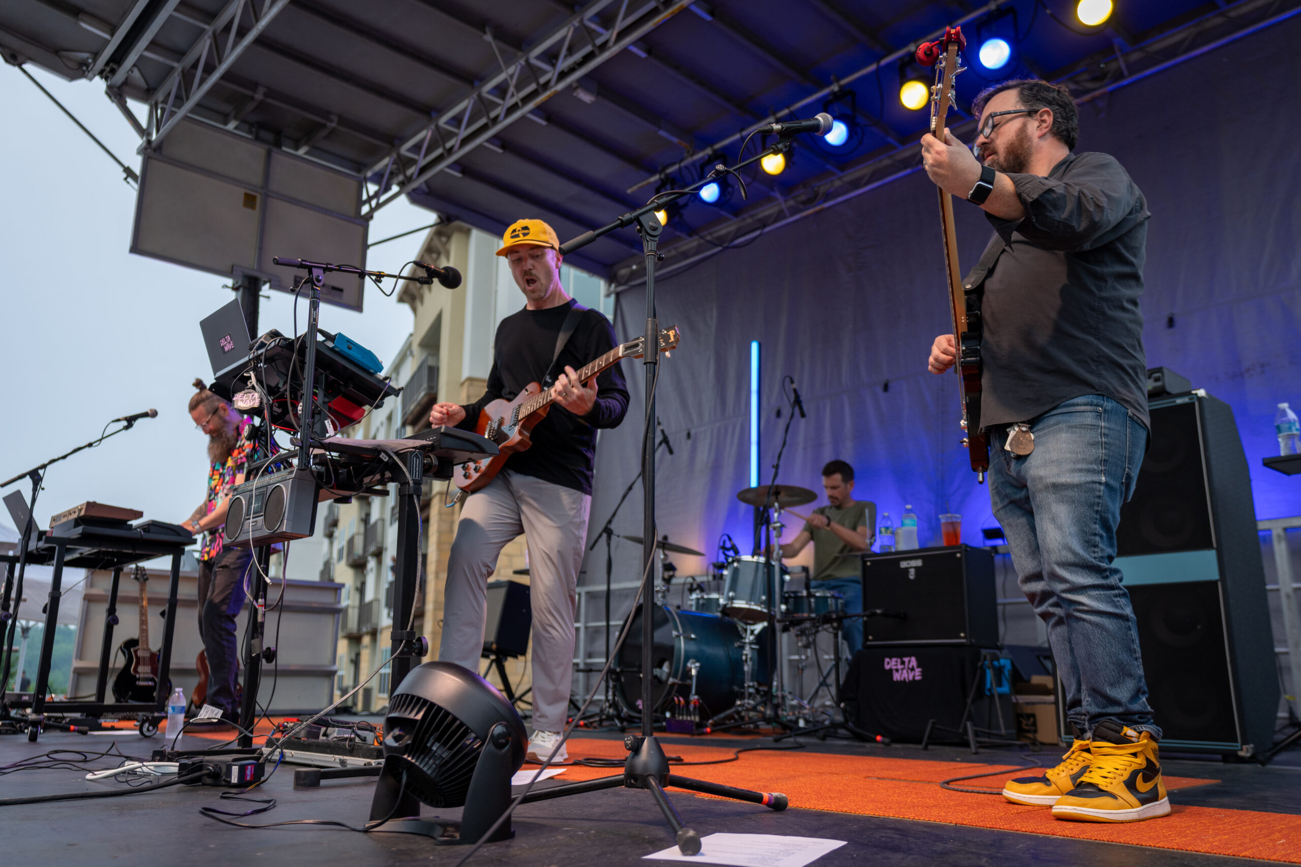 A four-member band performs on an outdoor stage at Canonsburg Live Music. One plays keyboard, another sings and plays guitar, the third plays bass, and the fourth is on drums. Colorful stage lights illuminate them as they play.