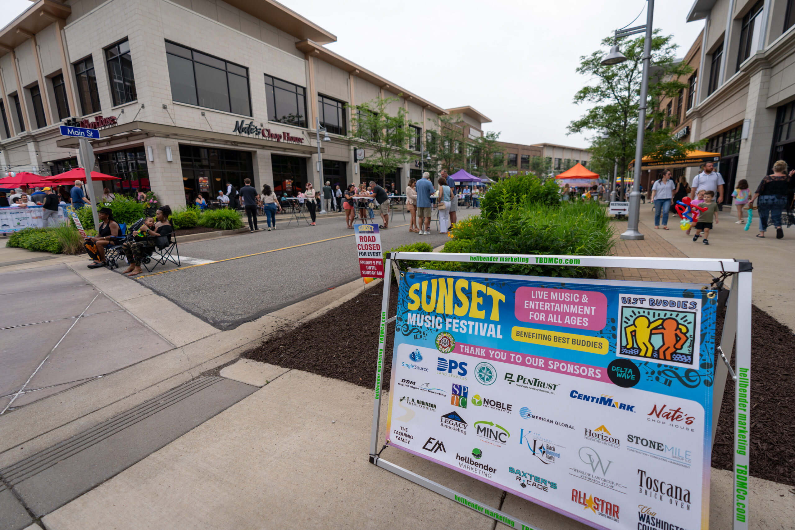 A colorful sign for the Sunset Music Fest stands in front of a street, where people gather near tents and shops for the outdoor event. The festival features Canonsburg live music and entertainment for all ages.