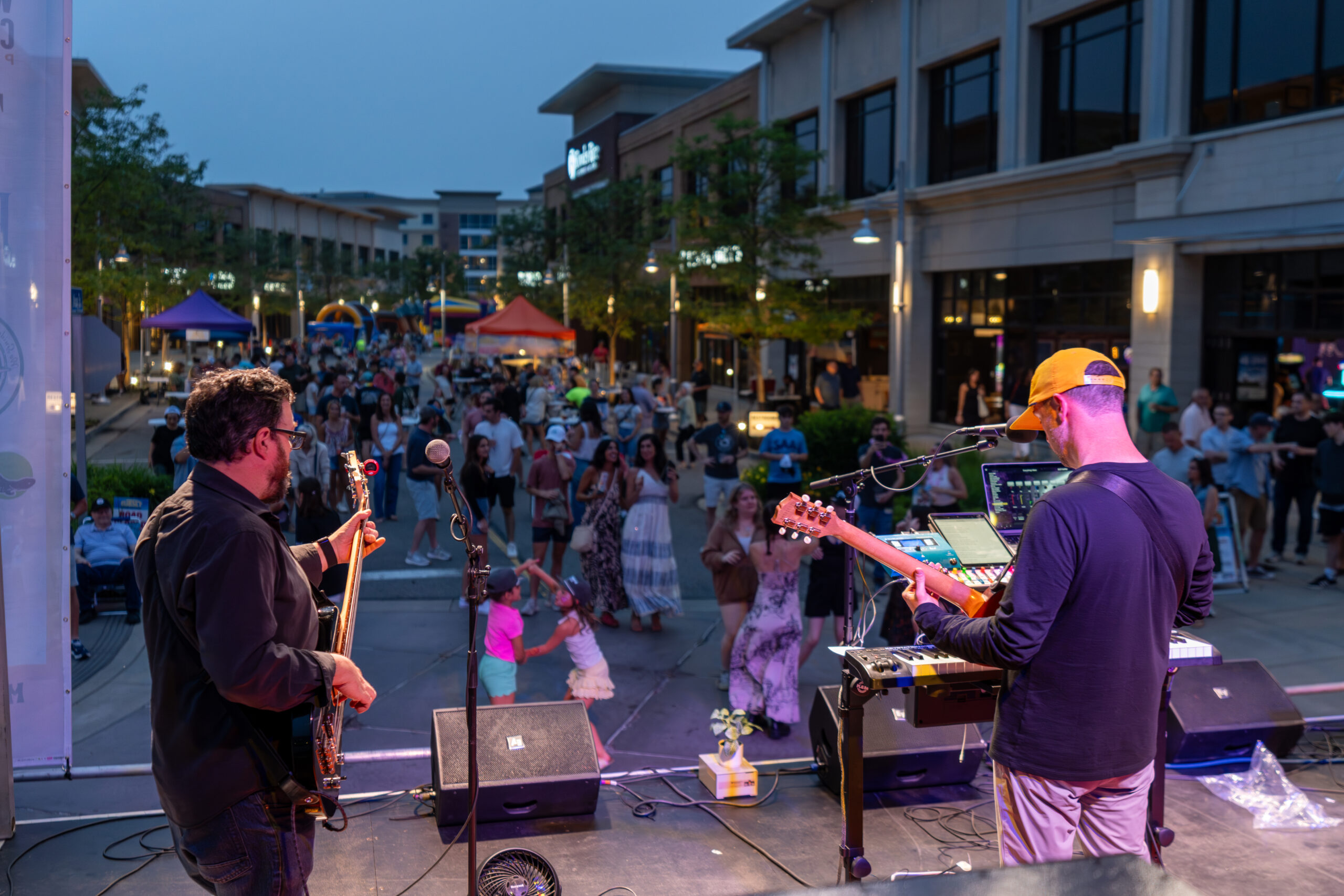 View from behind two musicians playing guitars on an outdoor stage at dusk, facing a lively crowd between modern buildings at the Sunset Music Fest, a vibrant Canonsburg live music event.