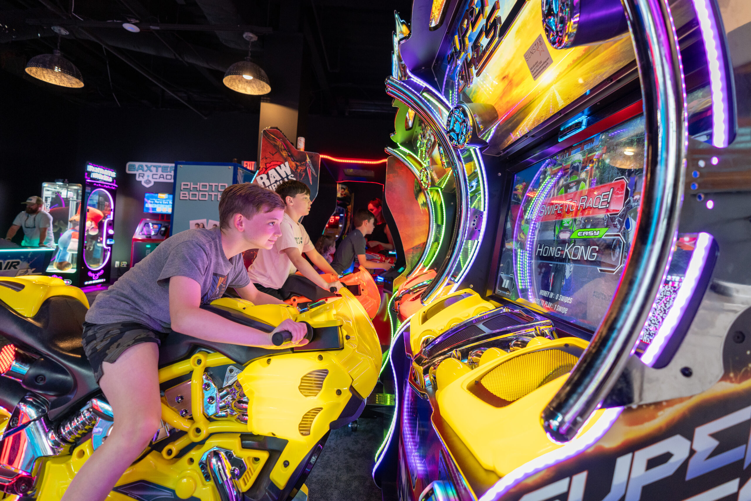 Two boys play a futuristic motorcycle racing arcade game, each riding bright yellow bike-shaped seats, surrounded by colorful lights and arcade machines in a lively game center during the Sunset Music Fest.