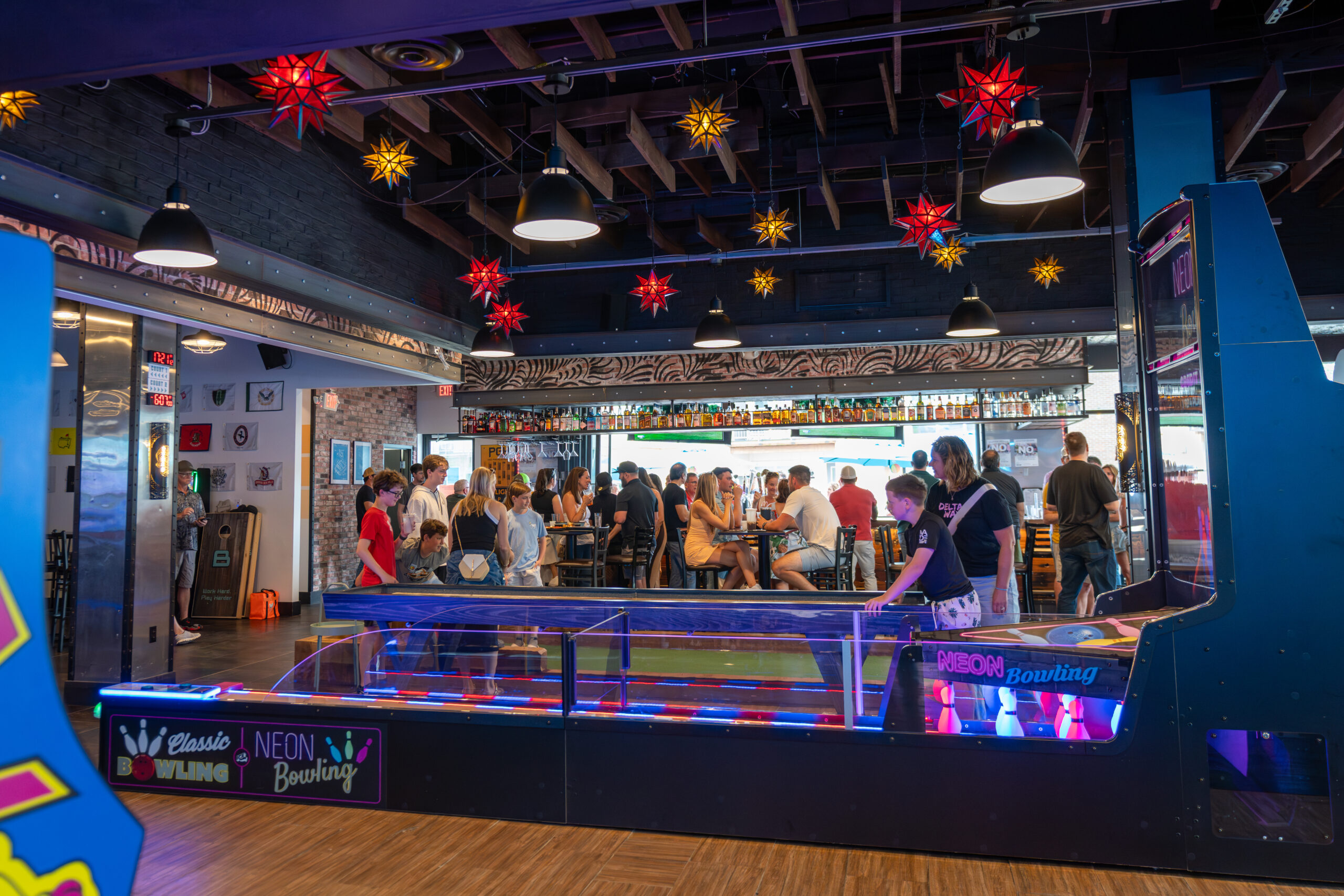 A lively indoor arcade scene shows people playing a neon-lit bowling game in the foreground, while groups socialize at tables under colorful star-shaped lights—capturing the vibrant spirit of Canonsburg Live Music events.