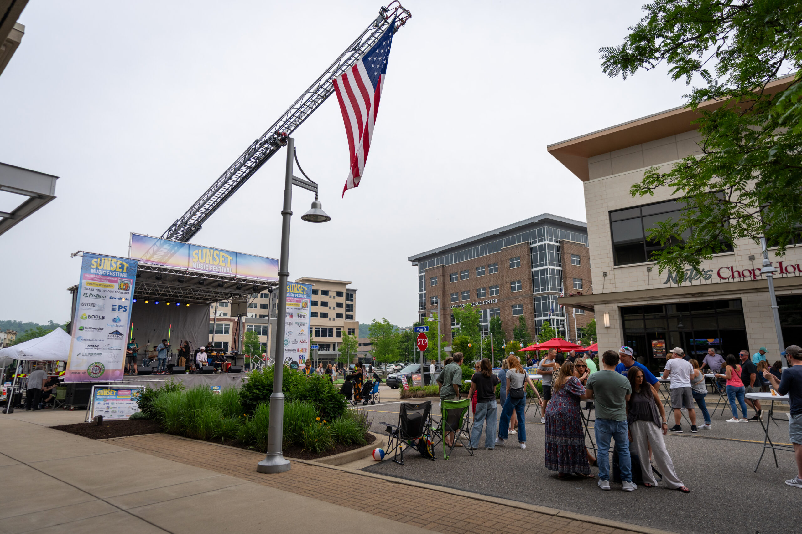 People gather on a closed-off street for the Canonsburg Live Music Festival, enjoying an outdoor concert on a banner-adorned stage as a large American flag hangs from a crane. Buildings and greenery surround the festive scene.