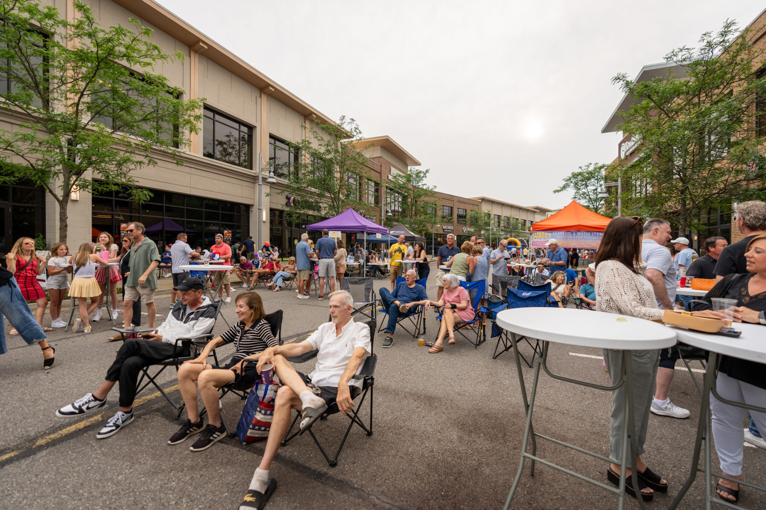 People gather at a lively outdoor street event, part of the Sunset Music Fest, with tents, tables, and chairs. Some are seated while others stand and socialize. Buildings and trees line the street under an overcast sky.