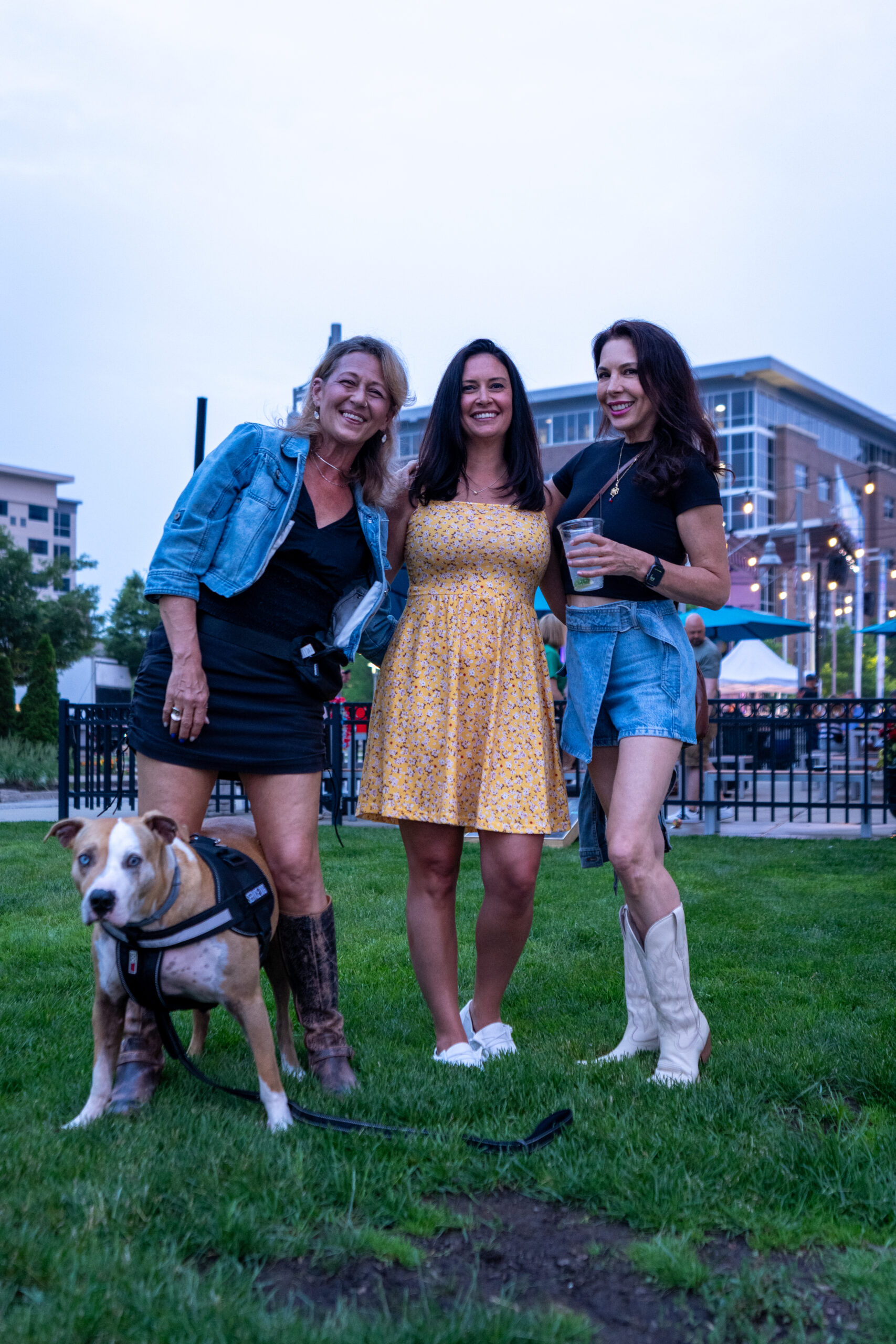 Three women stand together smiling outdoors on grass at Sunset Music Fest, with one holding a drink. A tan and white dog in a harness is in front of them. Buildings and patio lights create a lively music festival backdrop.
