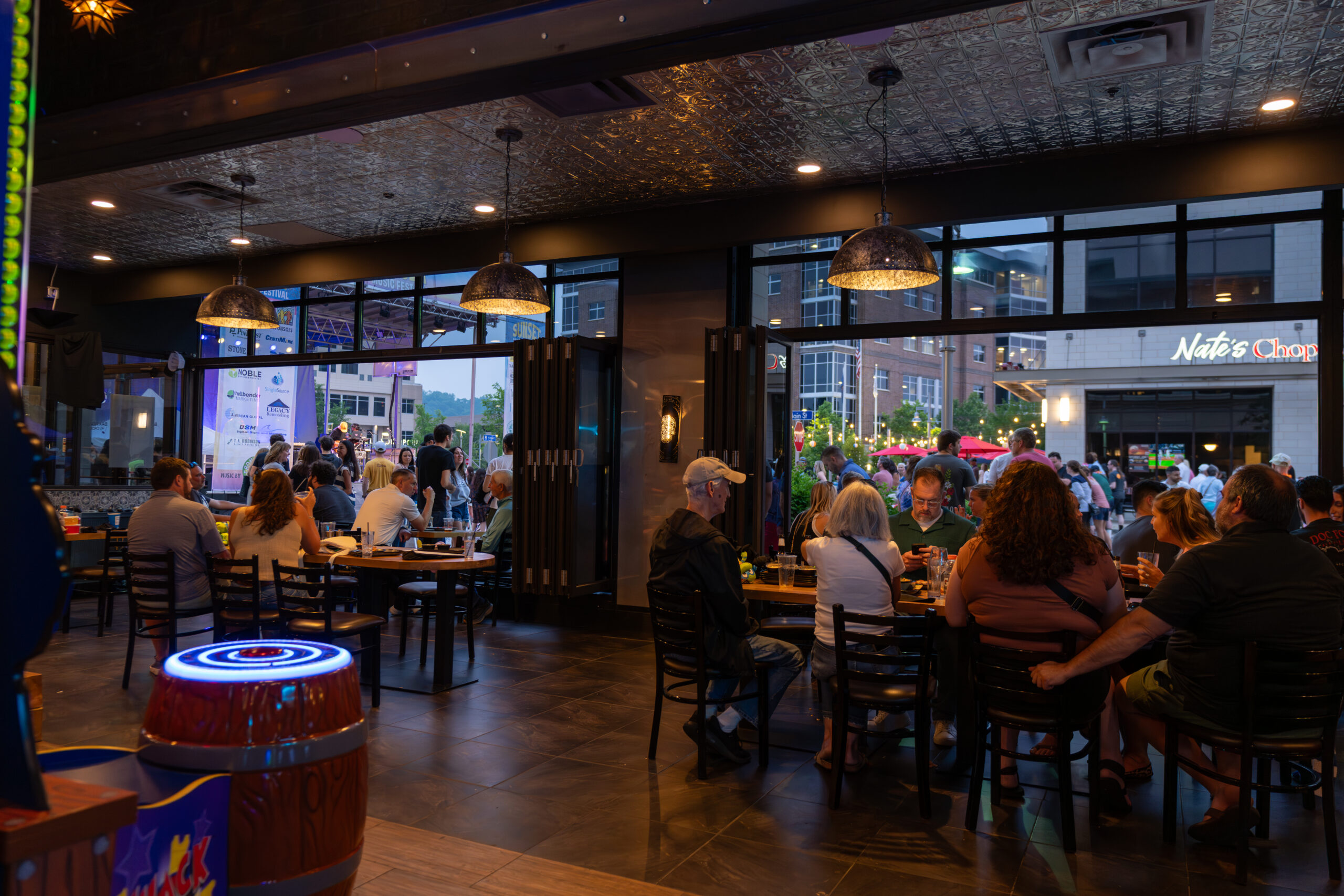 People are dining and socializing in a busy restaurant with large windows showing a lively outdoor scene and Nate’s Chop across the street. Warm lighting creates a cozy indoor atmosphere, perfect for discussing the upcoming Sunset Music Fest nearby.