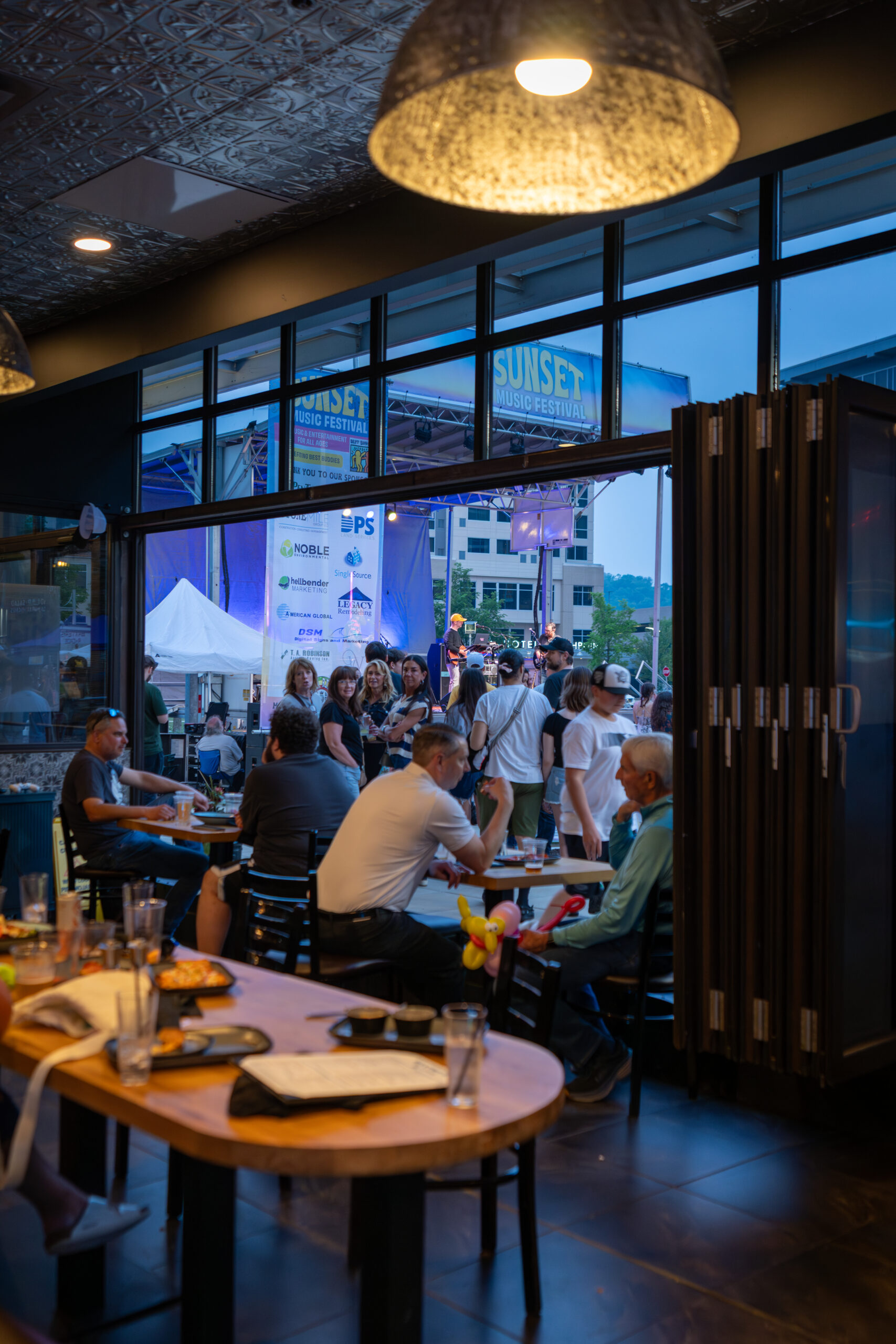 People dine inside a restaurant near large windows while a crowd gathers outside for the Sunset Music Fest, visible through the glass. Plates, glasses, and pizza are on tables in the foreground.