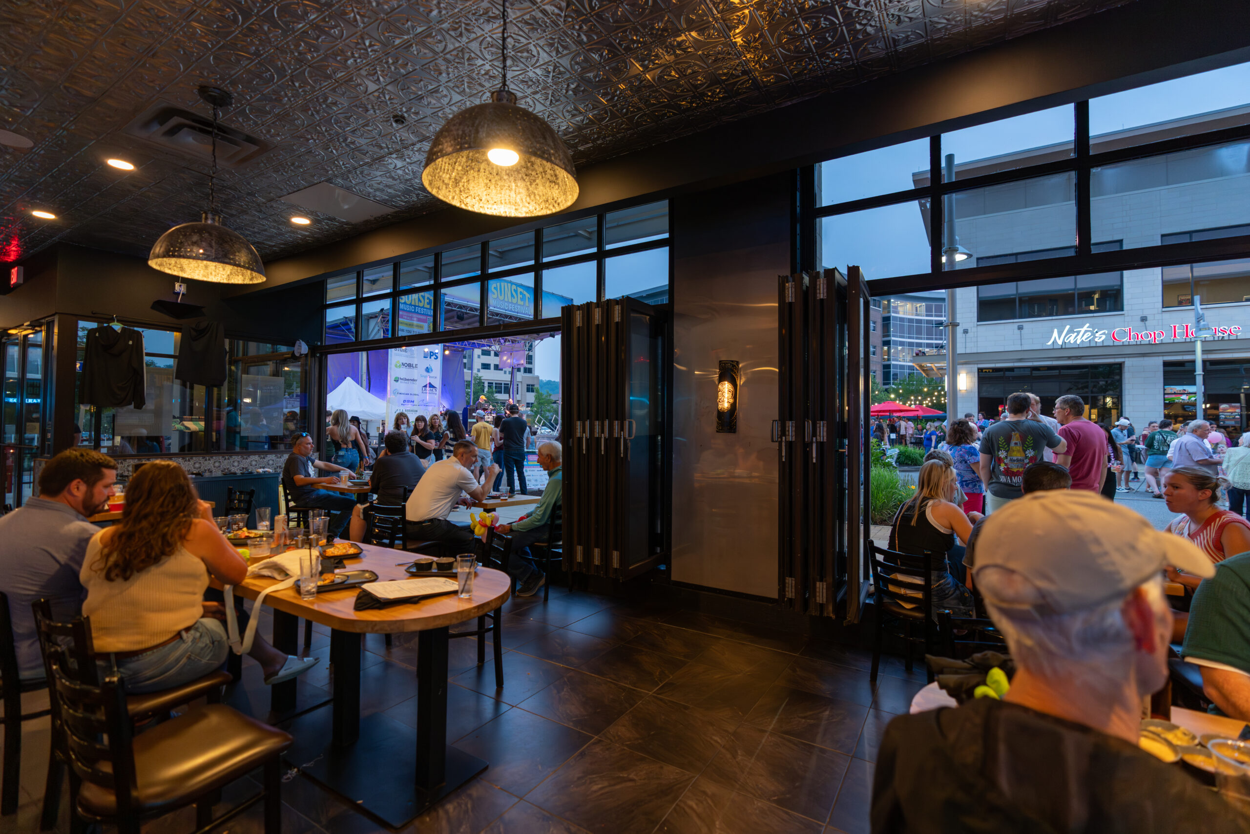 A busy restaurant scene at dusk, with diners seated at tables indoors and large windows revealing people gathered outside for the Canonsburg Live Music event. The atmosphere is lively and social.