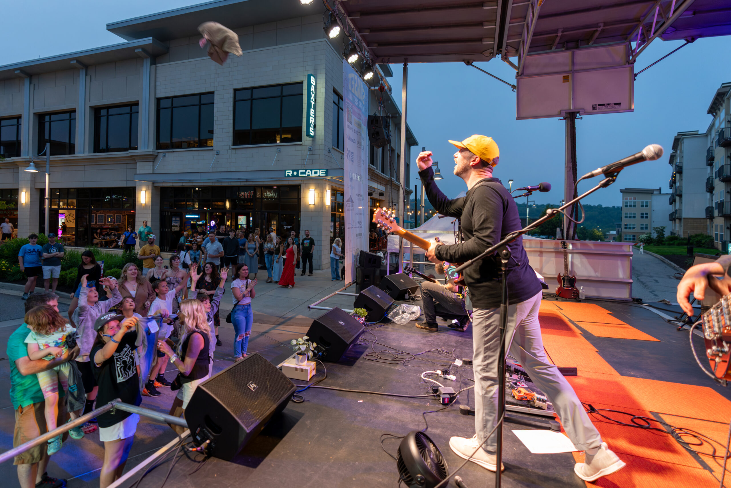 A musician on an outdoor stage tosses his hat to an enthusiastic crowd during a lively Canonsburg Live Music concert, with shops and onlookers visible in the background.