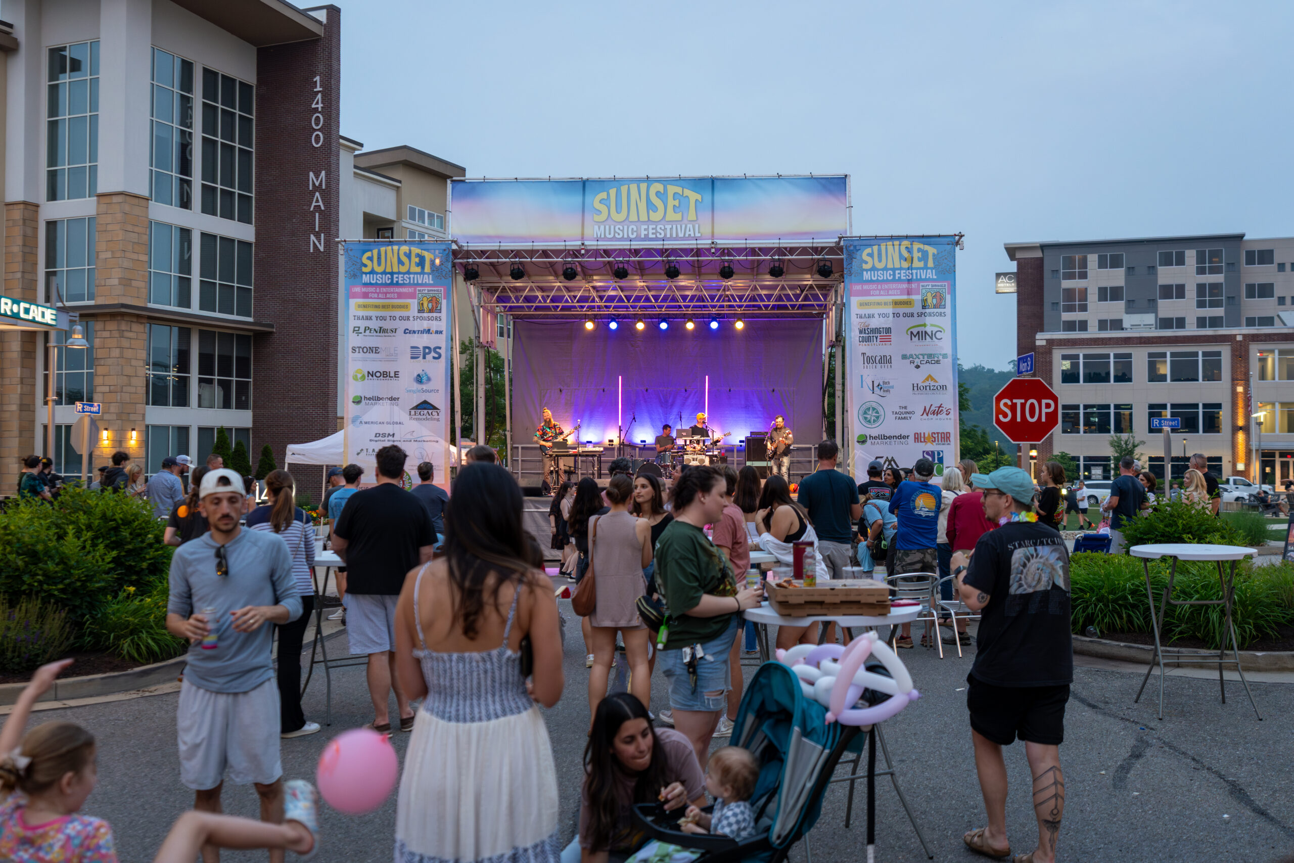 A crowd enjoys live music at the Sunset Music Fest, with people standing, sitting, and walking near food tables. Apartment buildings and banners frame the stage in the background of this lively Canonsburg live music event.